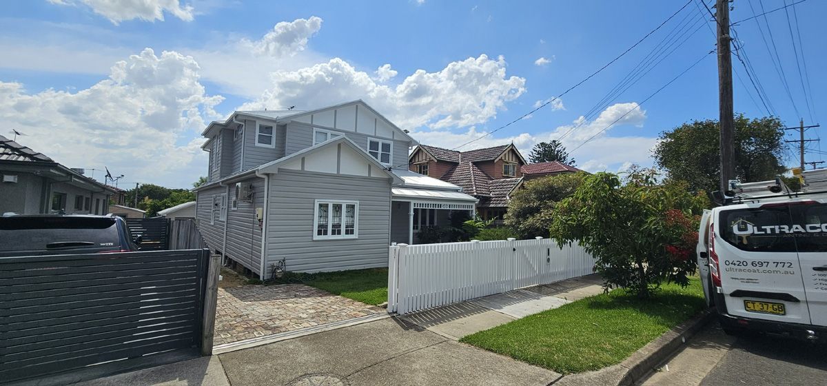 Small white house with a front fence on a sunny residential street, with a van parked nearby.
