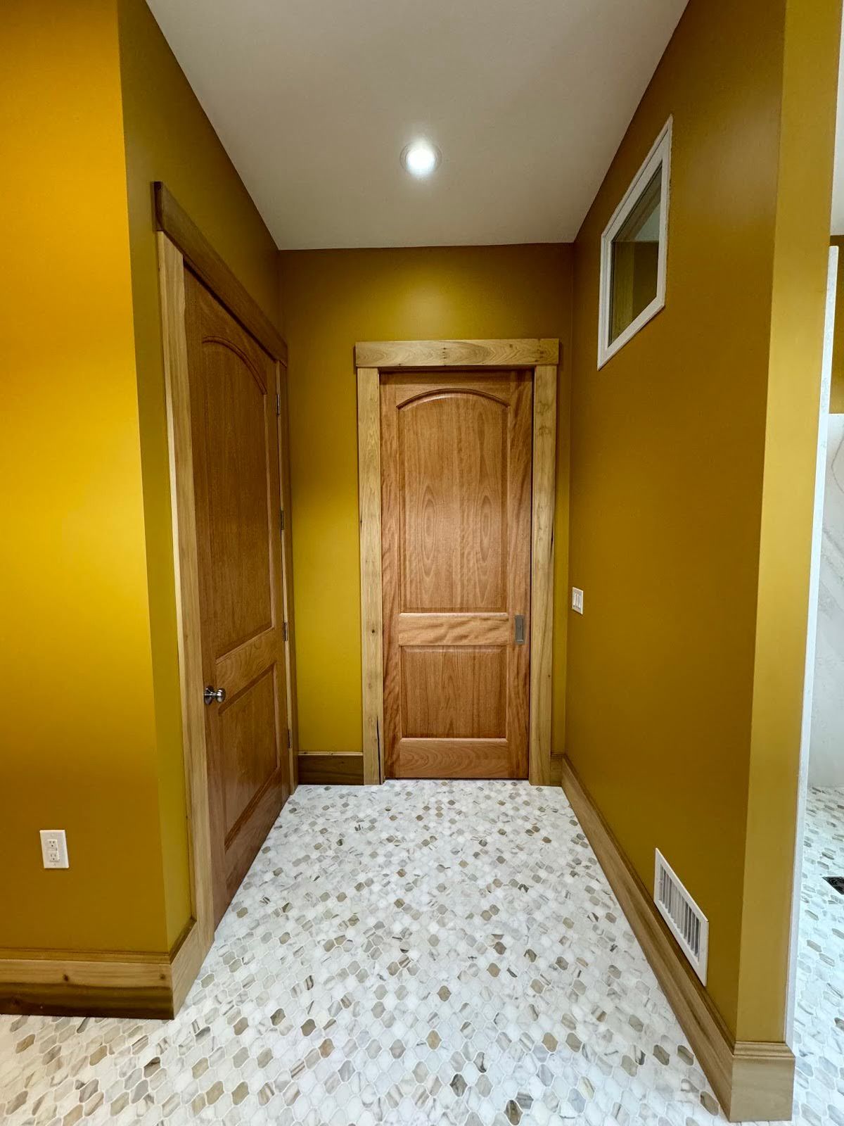 Narrow hallway with yellow walls, wooden doors, and patterned tile floor, lit by a ceiling light