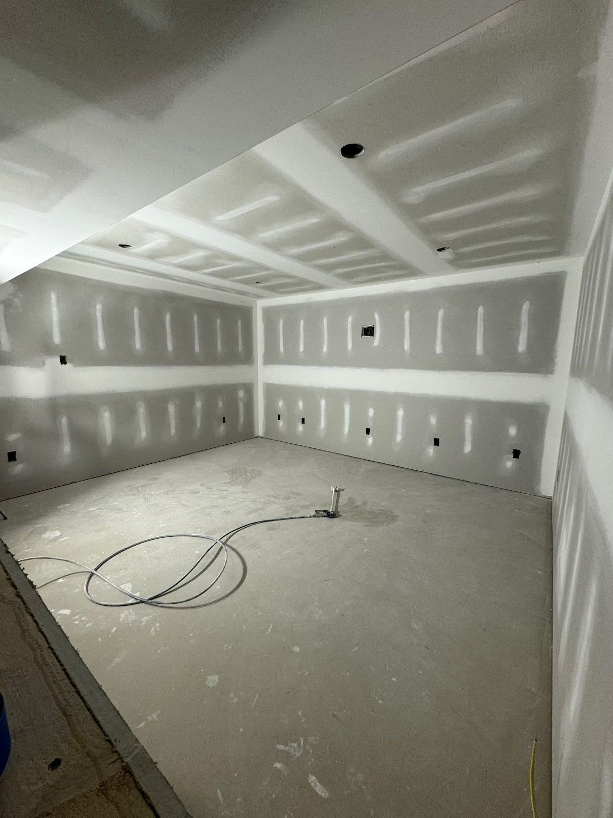 Unfinished drywall room with white walls, ceiling patches, and cables coiled on a dusty floor.
