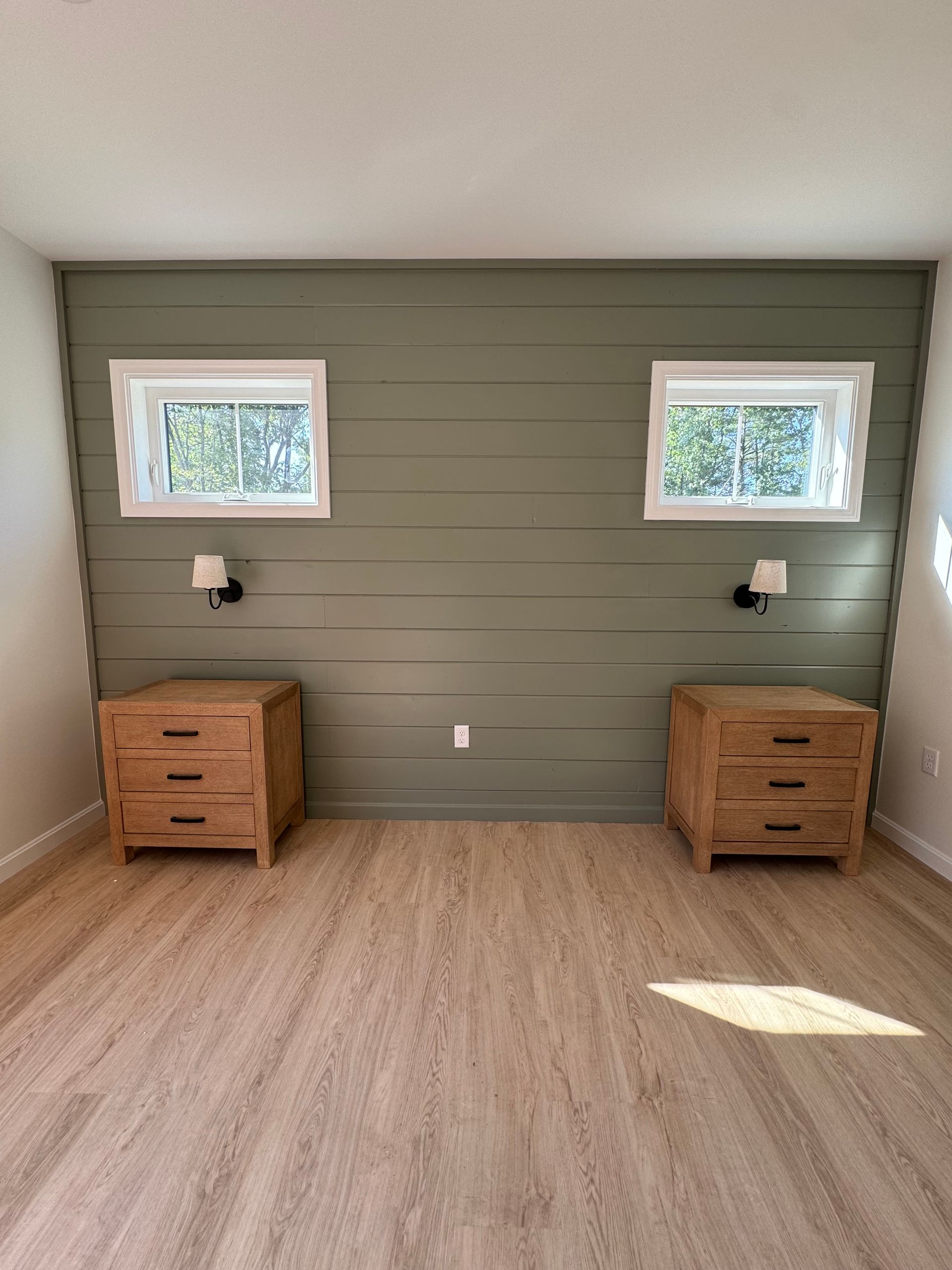 Empty room with sage green accent wall, two small windows, and two wooden nightstands on a light wood floor.
