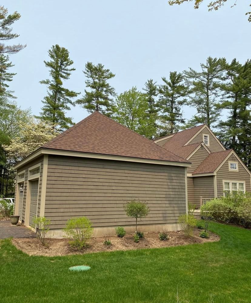 Tan house with brown shingle roof beside a grassy yard and tall trees under a clear sky
