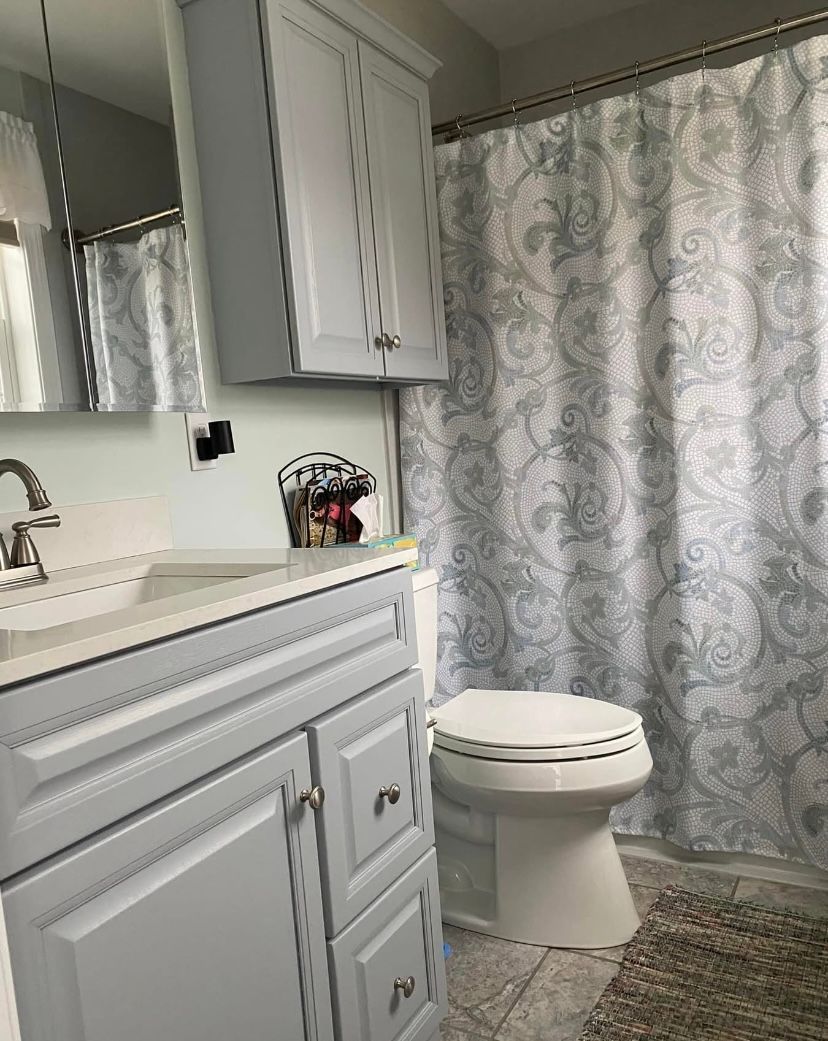 White bathroom vanity and toilet beside a gray patterned shower curtain.