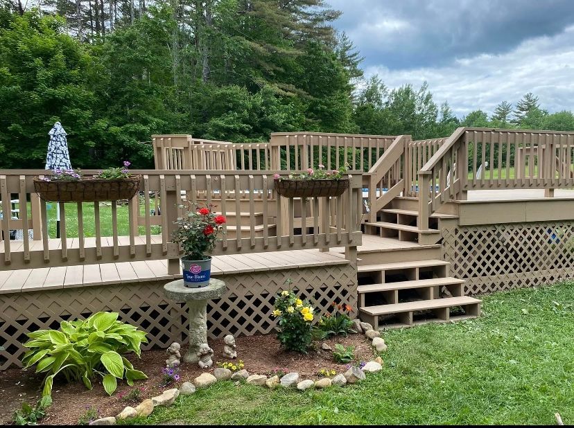 Wooden backyard deck with steps, railing, potted flowers, and lattice skirting beside a grassy lawn and trees