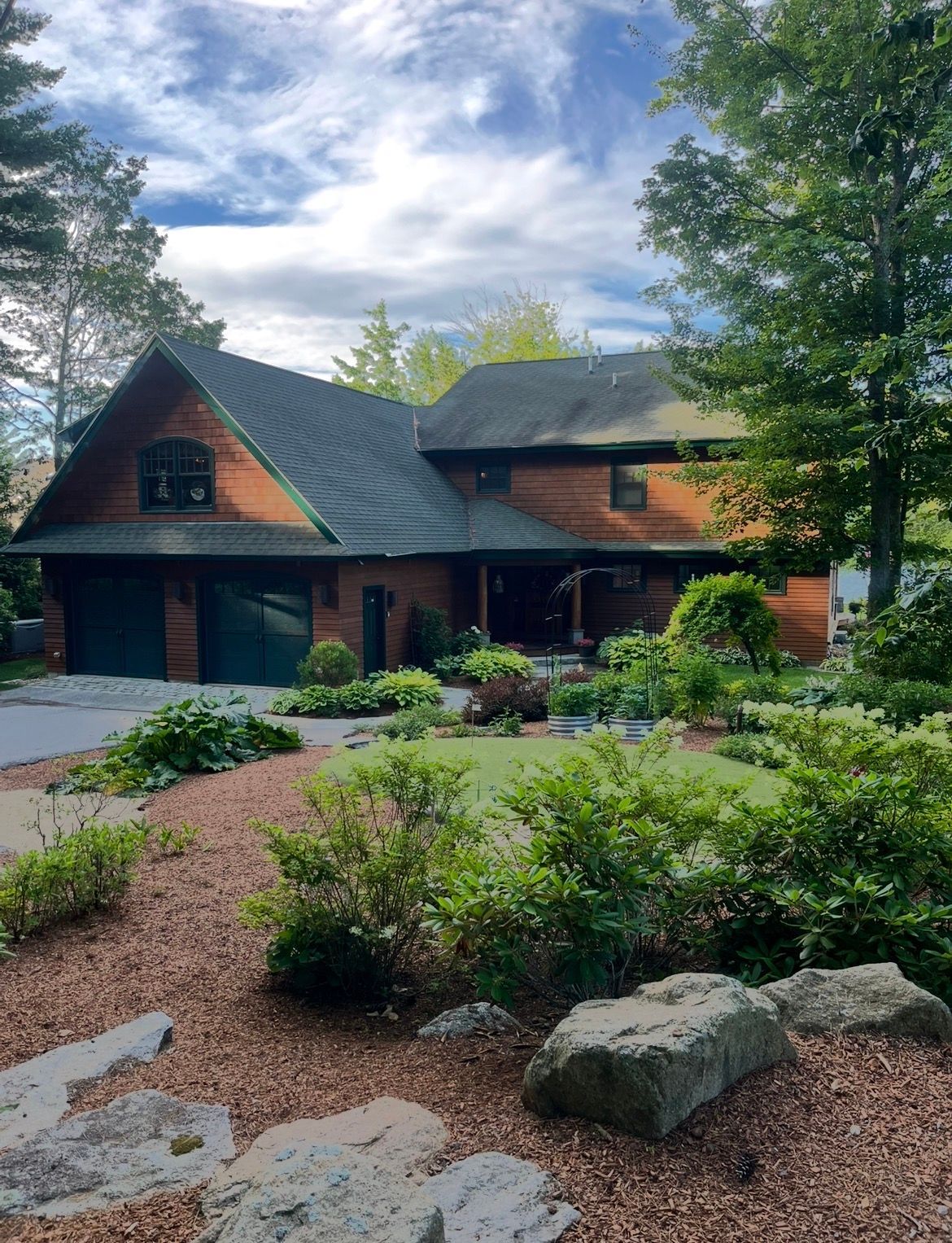 Brick house with dark roof behind a landscaped garden and trees under a cloudy sky