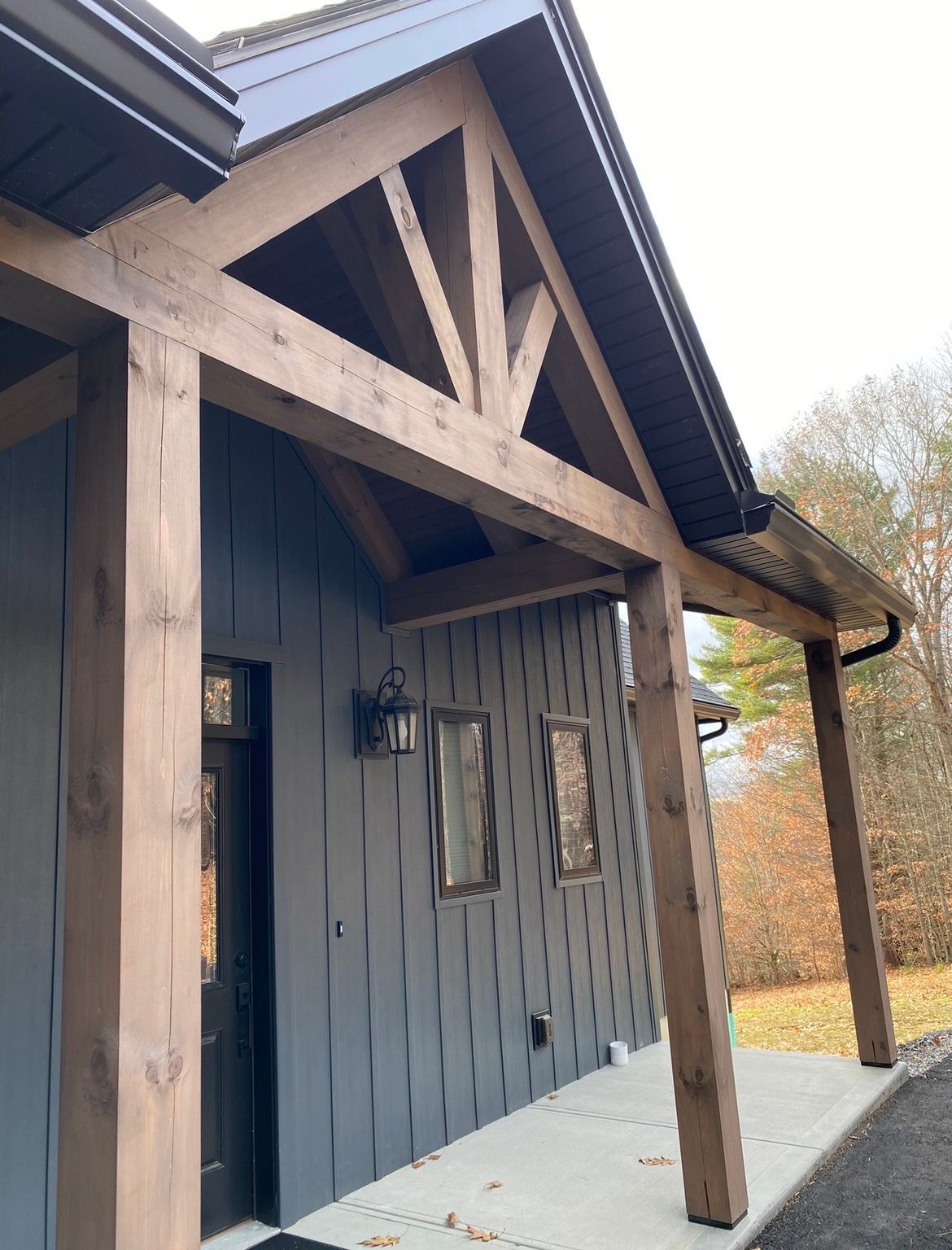 Covered wooden porch with dark gray siding and exposed timber beams, beside a gravel path in autumn.