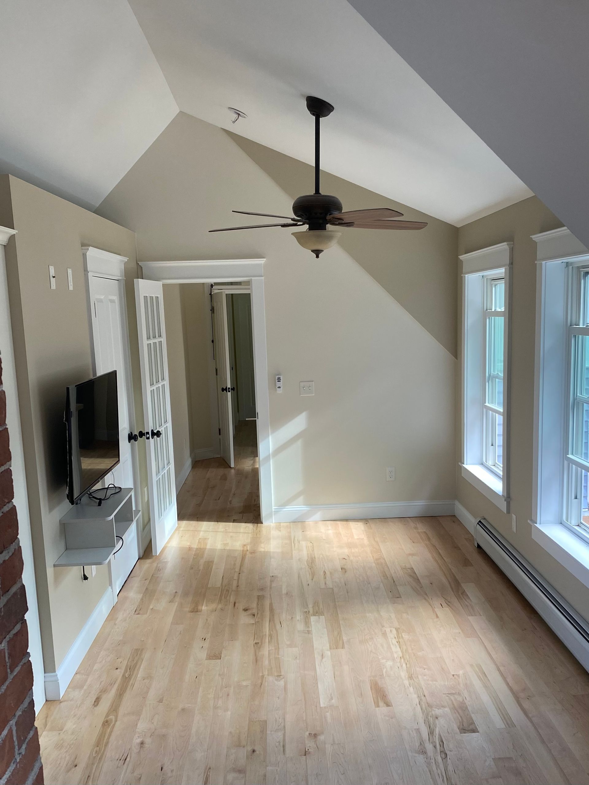 Empty sunlit room with hardwood floors, vaulted ceiling, ceiling fan, TV, and windows along one wall