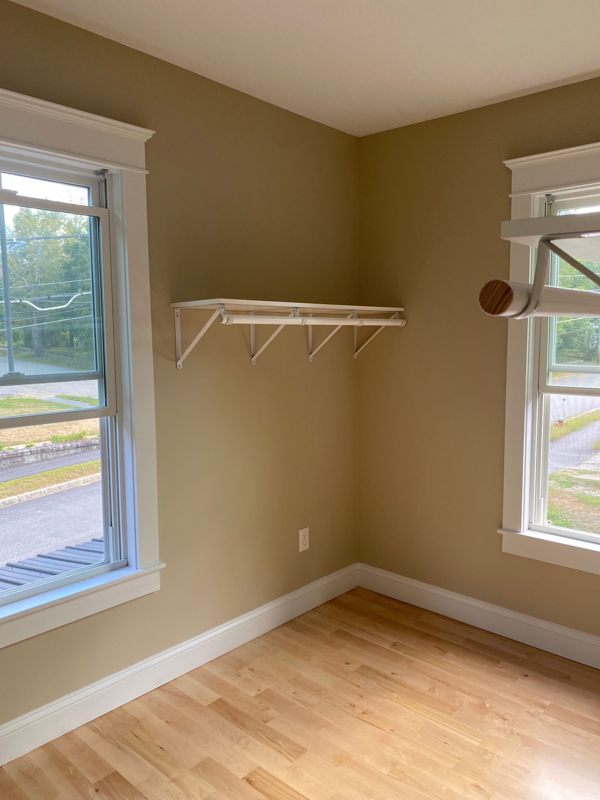 Empty corner of a room with beige walls, wood floor, two windows, and a white wall shelf.