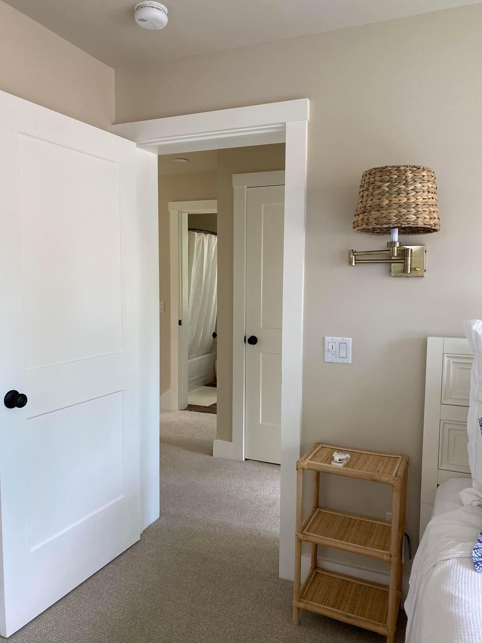 Bedroom doorway and hallway with white doors, beige carpet, wall lamp, and a small wicker shelf.