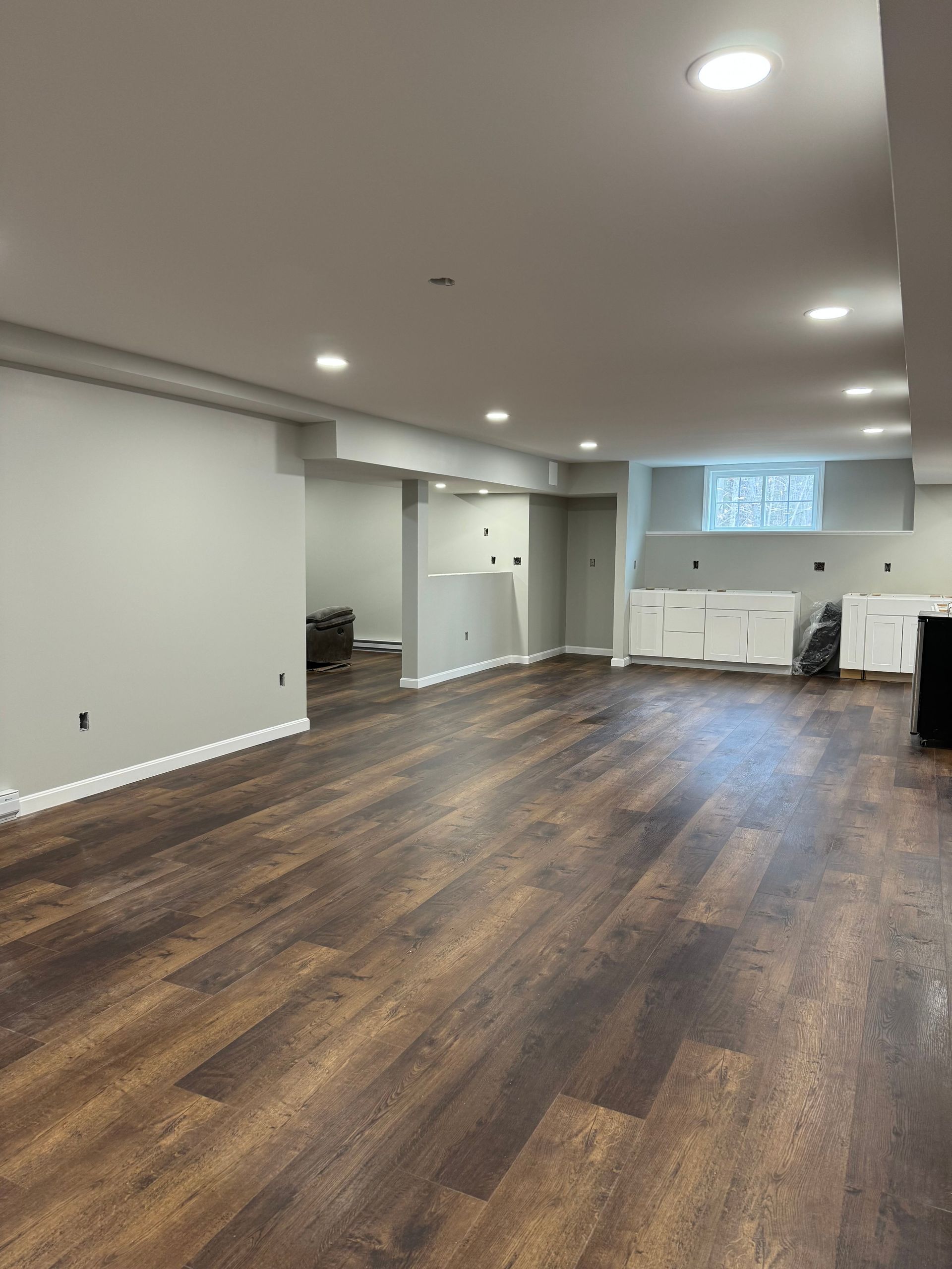 Empty finished basement with dark wood floors, white walls, recessed lights, and a small kitchen area.