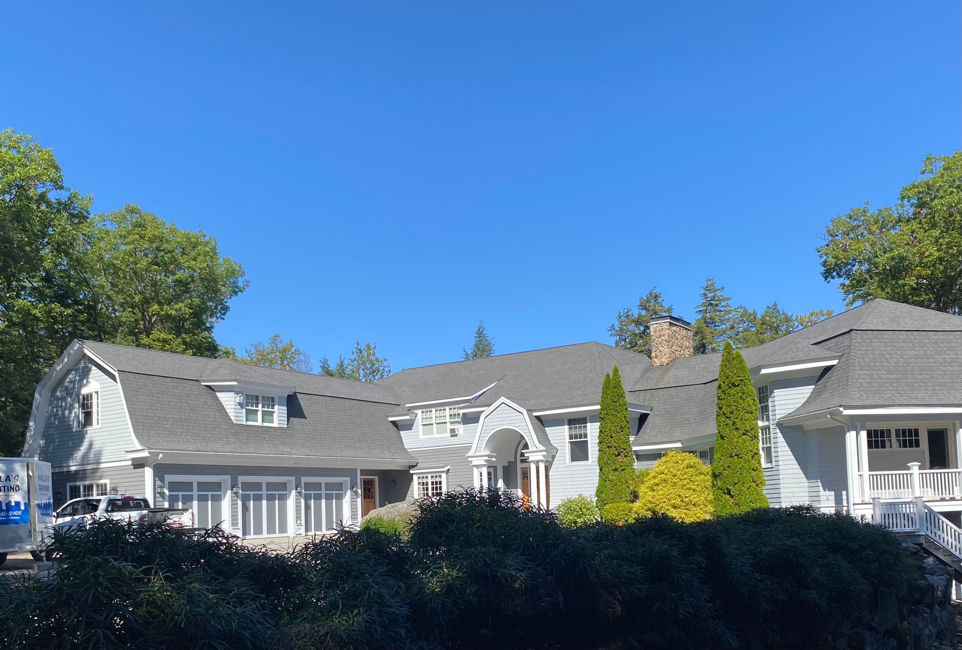 Suburban house with gray roof, white trim, and lush trees under a clear blue sky