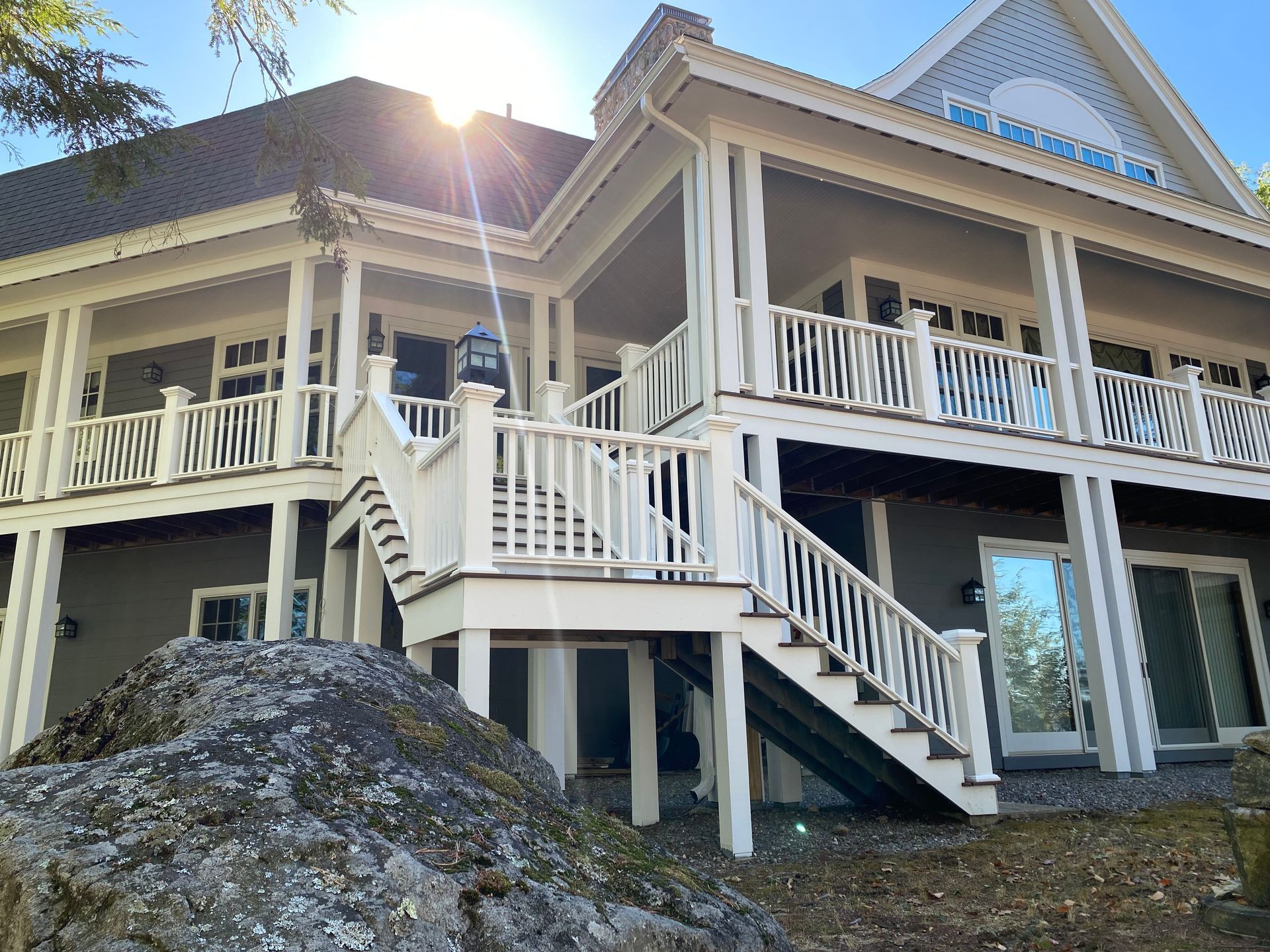 Two-story gray house with white trim, wraparound porch, and stairs on a rocky hillside in sunlight