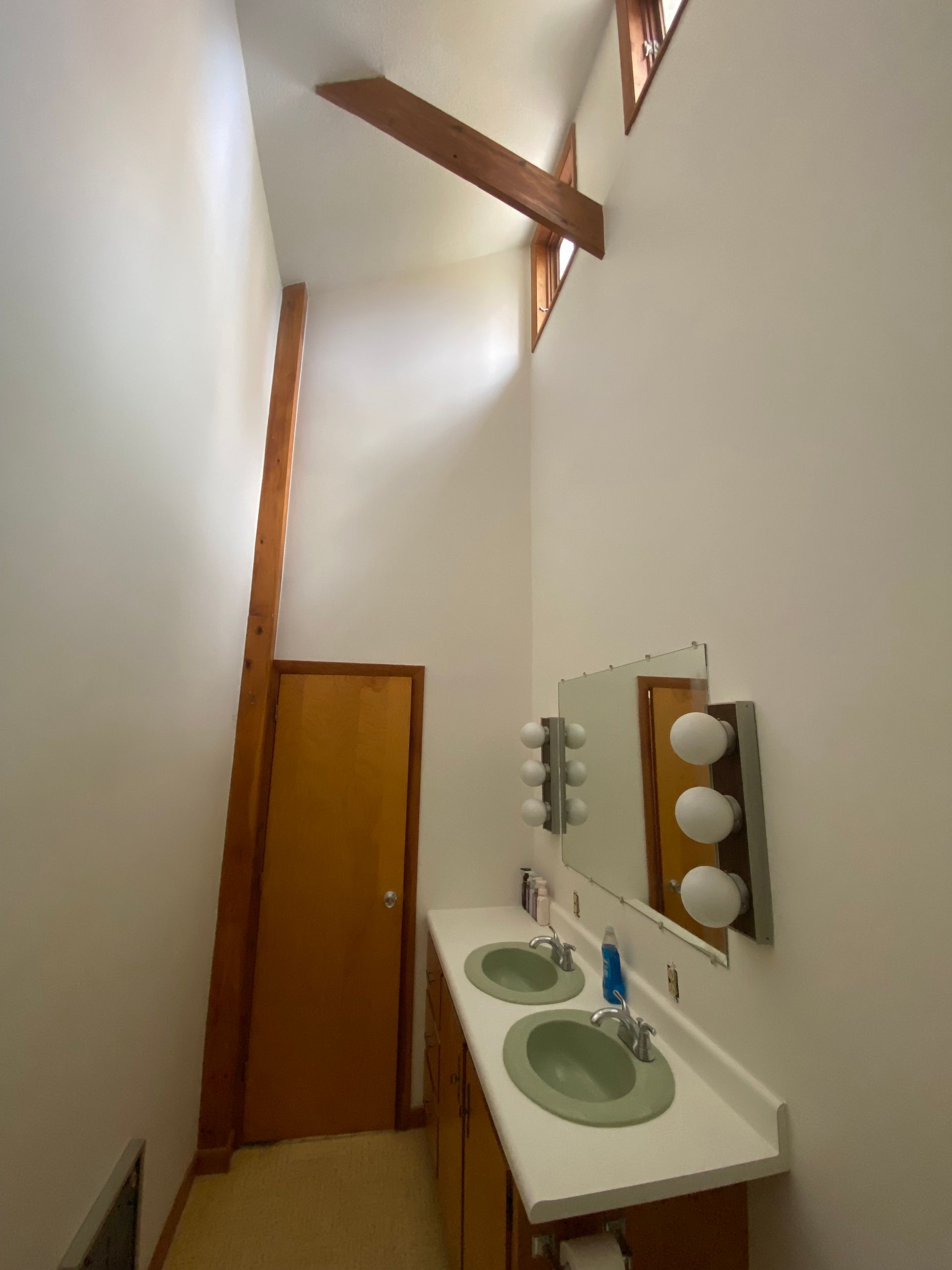 Narrow bathroom with two green sinks, wall mirror, wooden door, and high angled ceiling beams.