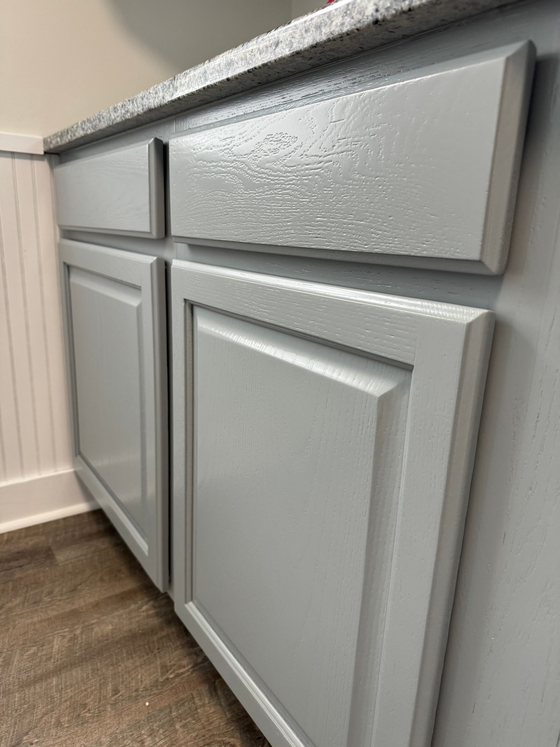 Gray kitchen cabinets with silver knobs and a wood floor, viewed at an angle.