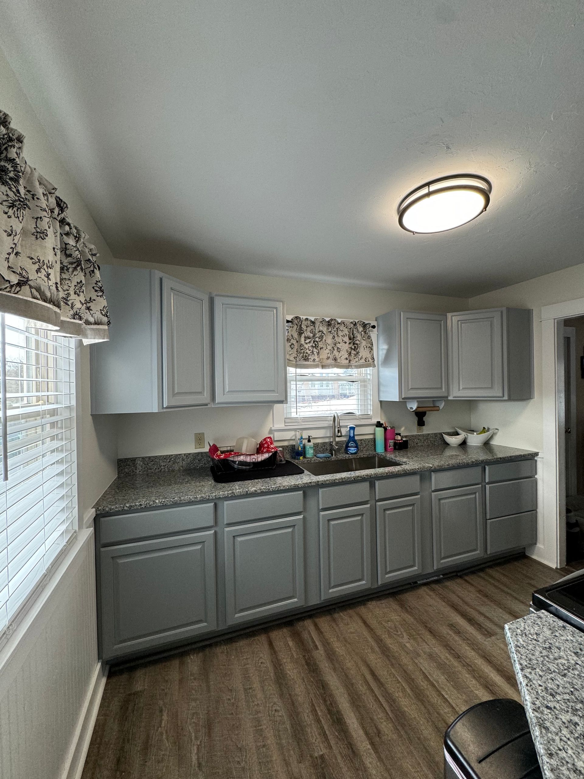 Small kitchen with gray cabinets, wood-look floor, white blinds, and a round ceiling light.