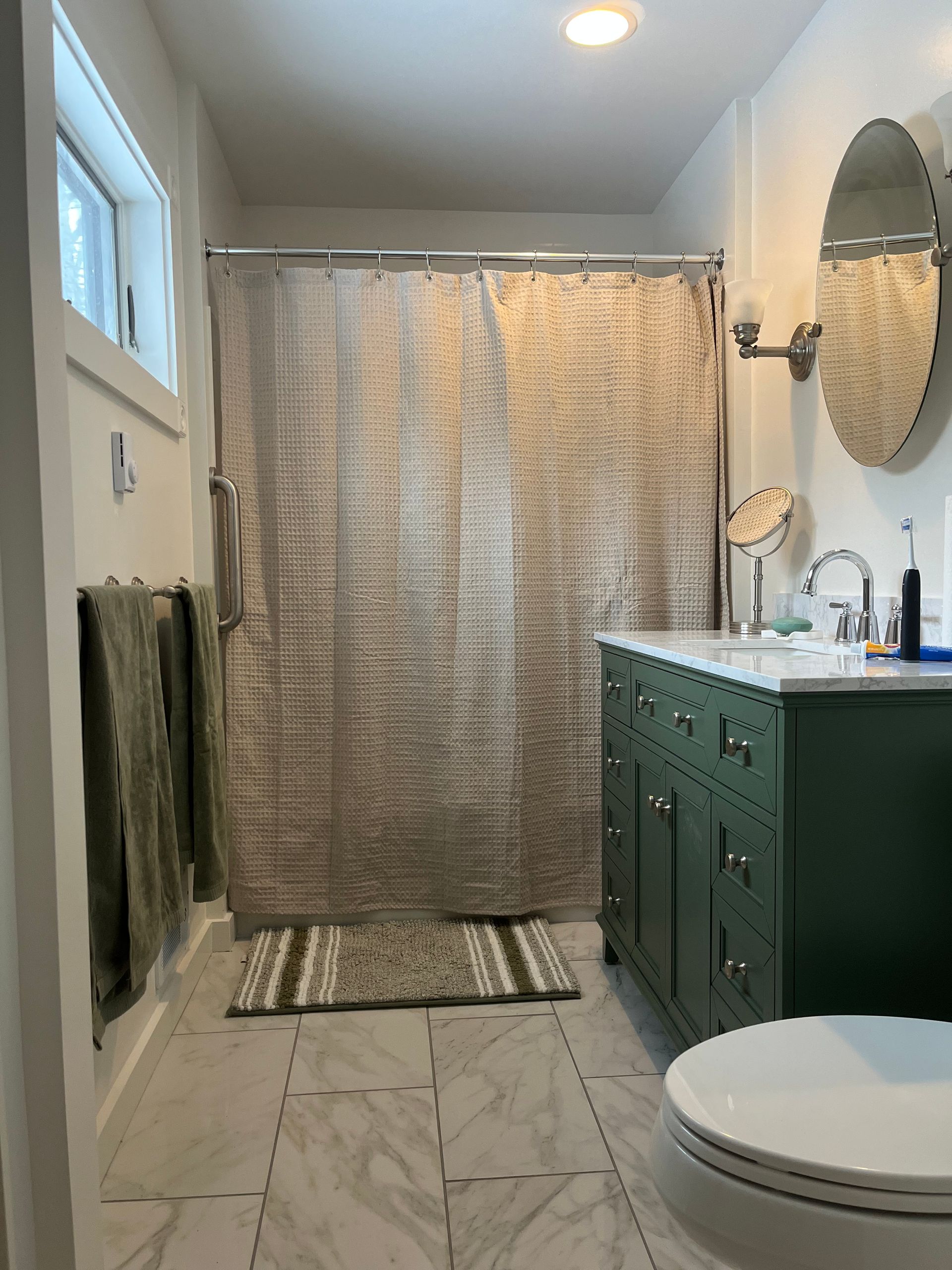 Narrow bathroom with green vanity, oval mirror, shower curtain, and a toilet in the foreground.
