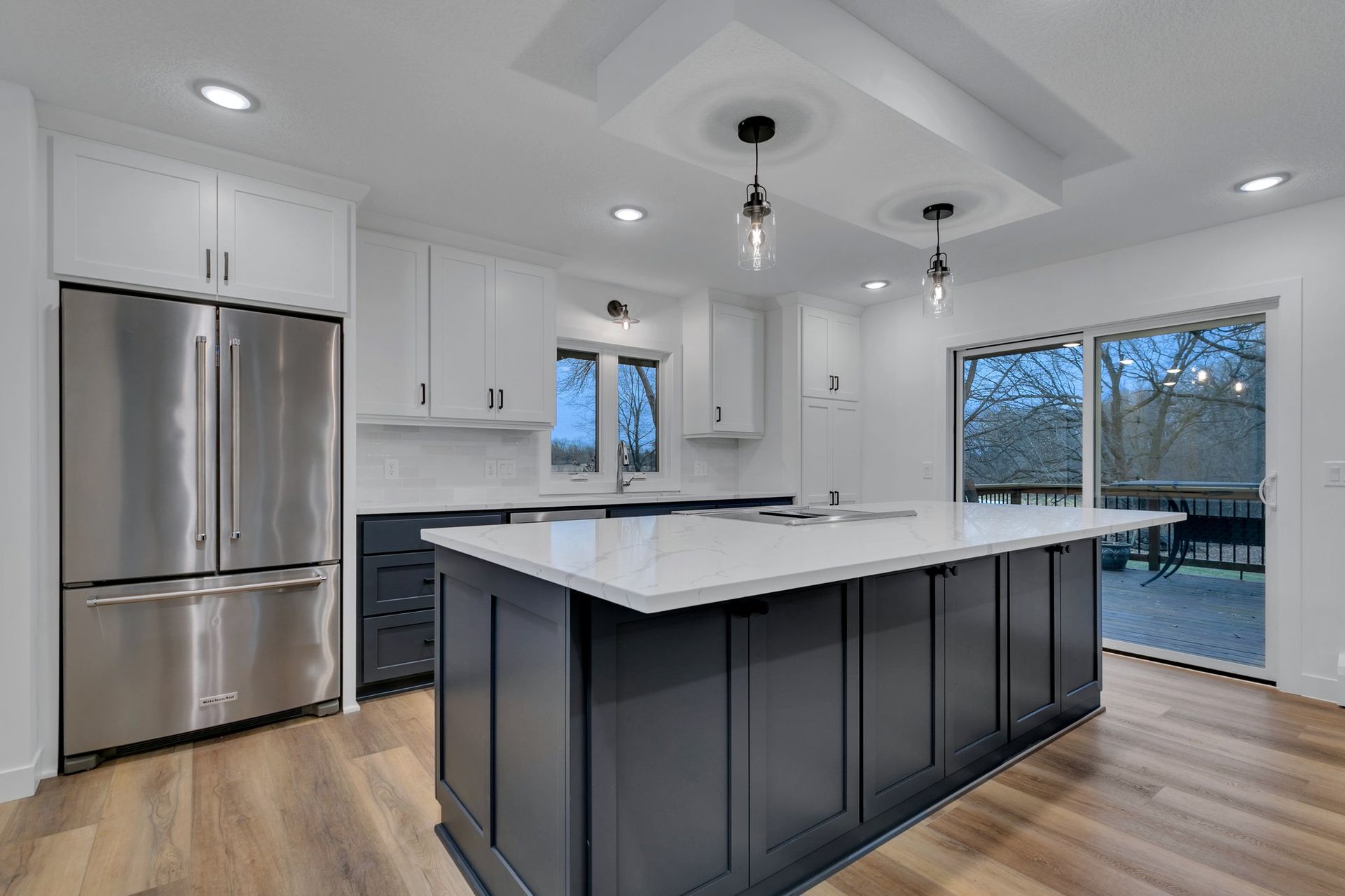 Bright modern kitchen with white cabinets, dark island, stainless fridge, and large windows overlooking outside