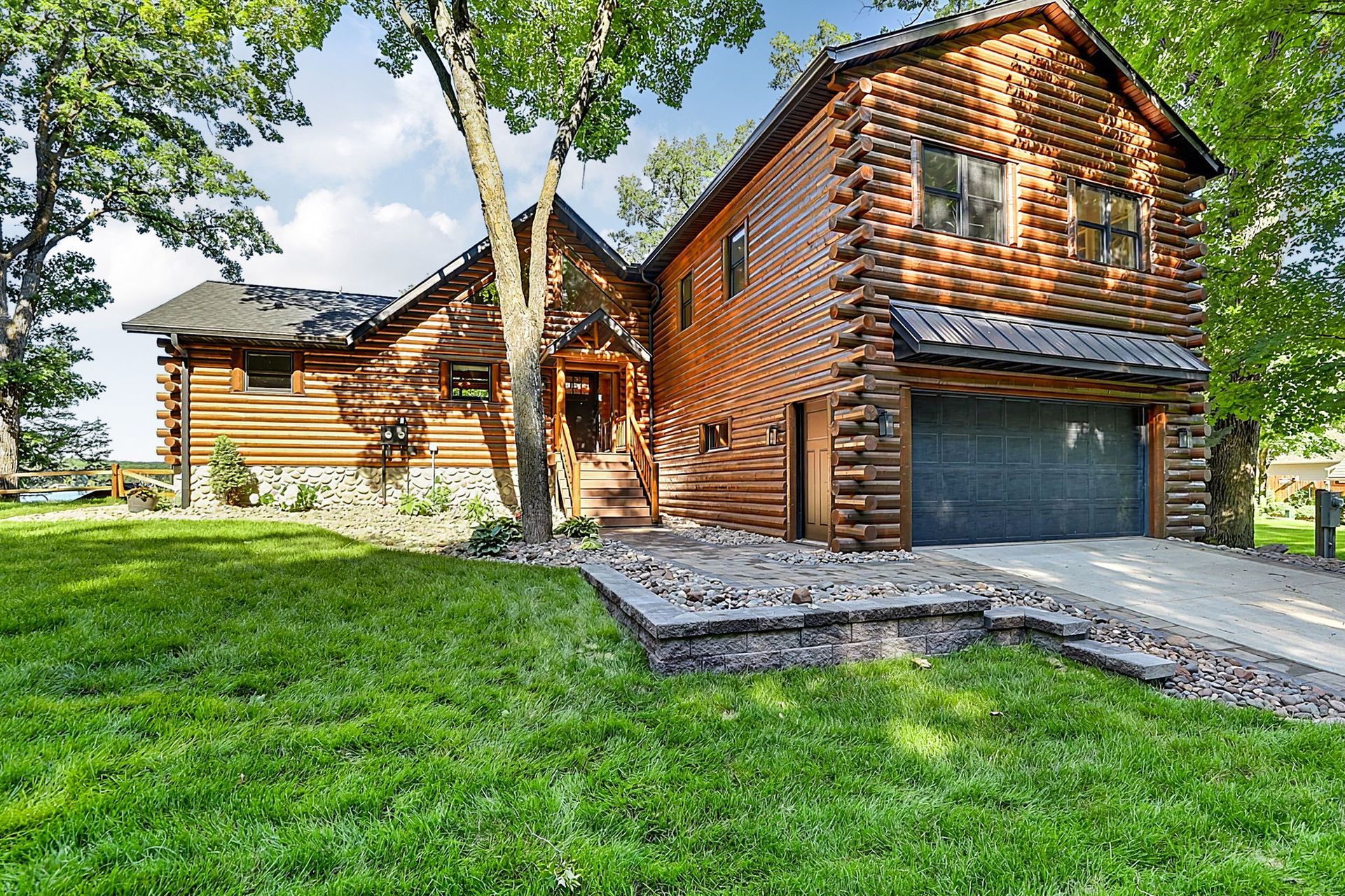 Wooden two-story house with a garage, gravel driveway, and green lawn under trees