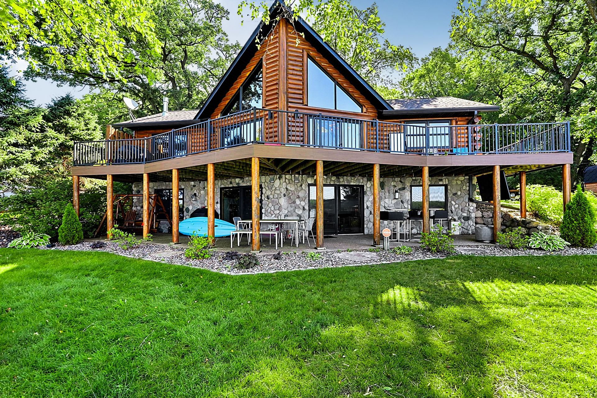 Wooden cabin with a wraparound deck in a lush green yard, surrounded by trees