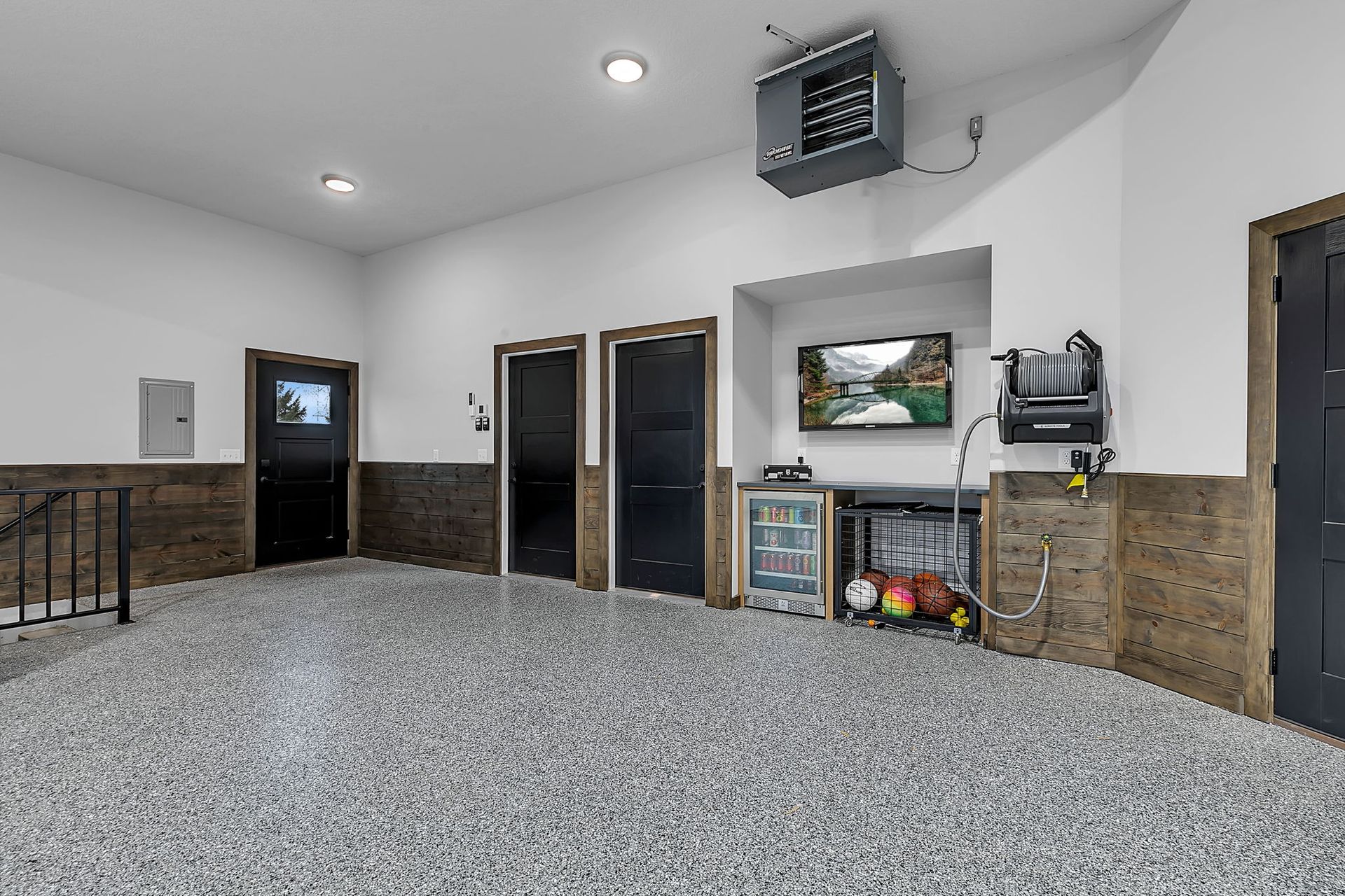 Garage interior with gray speckled floor, three black doors, TV, and heater. Walls are white and wood paneled.