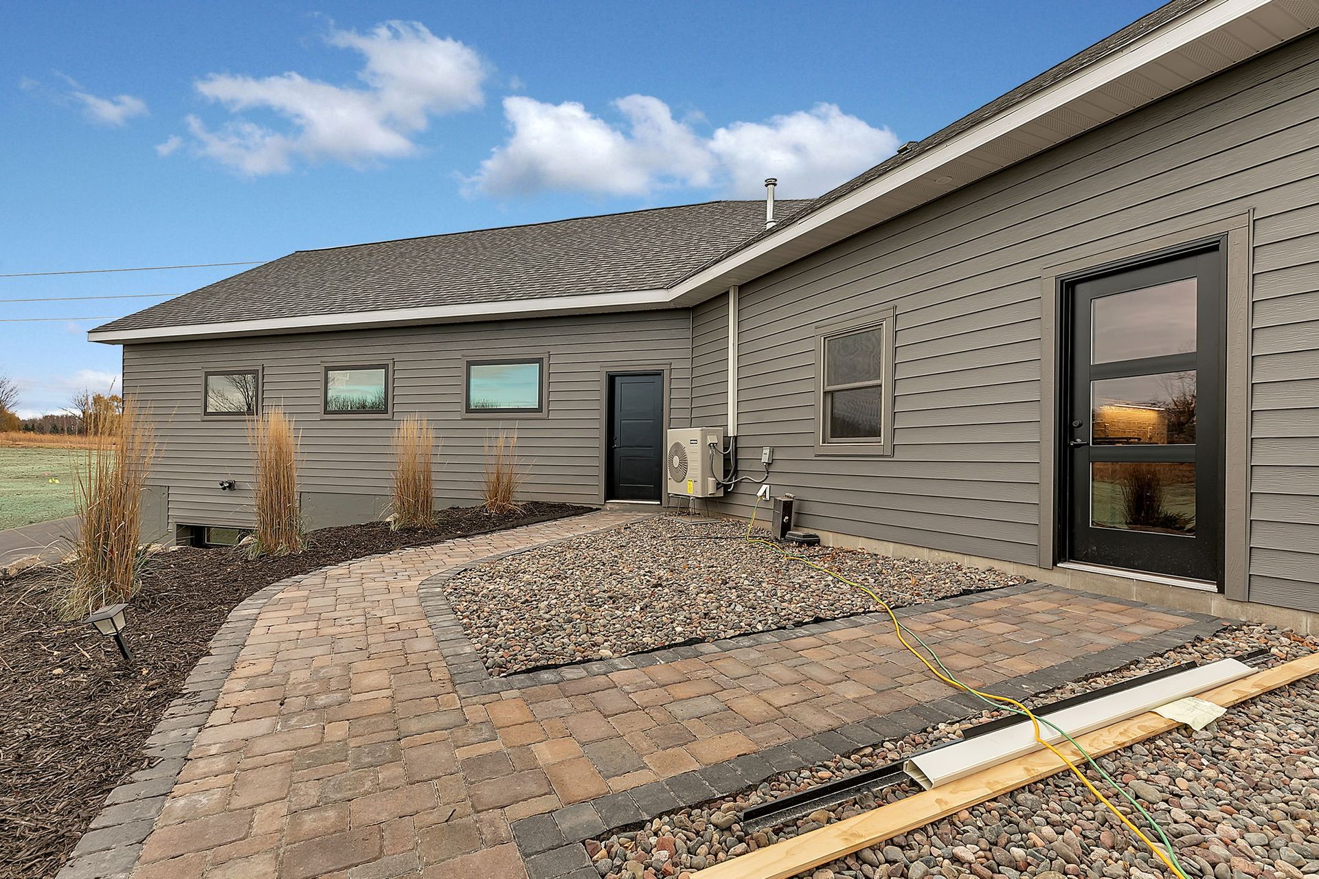 Gray house with dark door, patio, and landscaping under a blue sky.