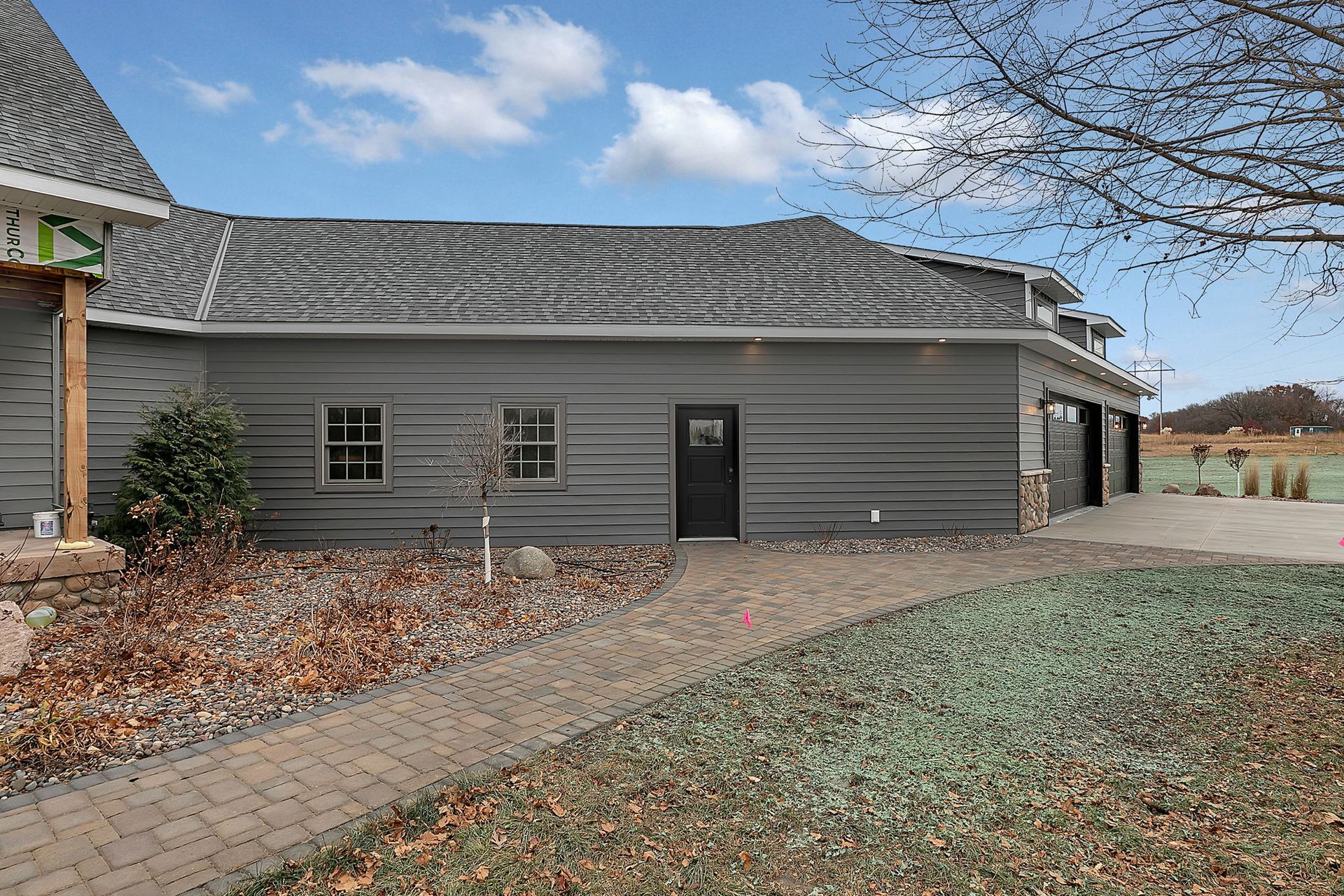Gray house with brick walkway, small windows, black door, and garage doors under a cloudy sky.