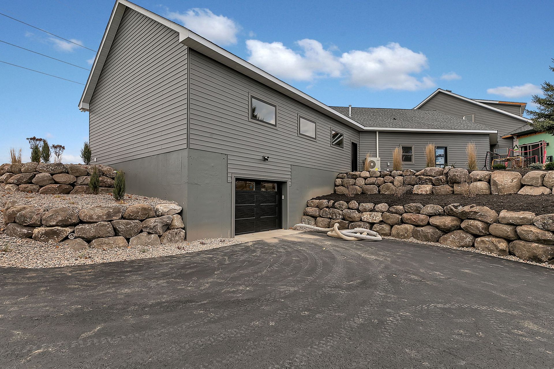 Gray building with a black garage door, set on a stone wall and driveway, under a blue sky.