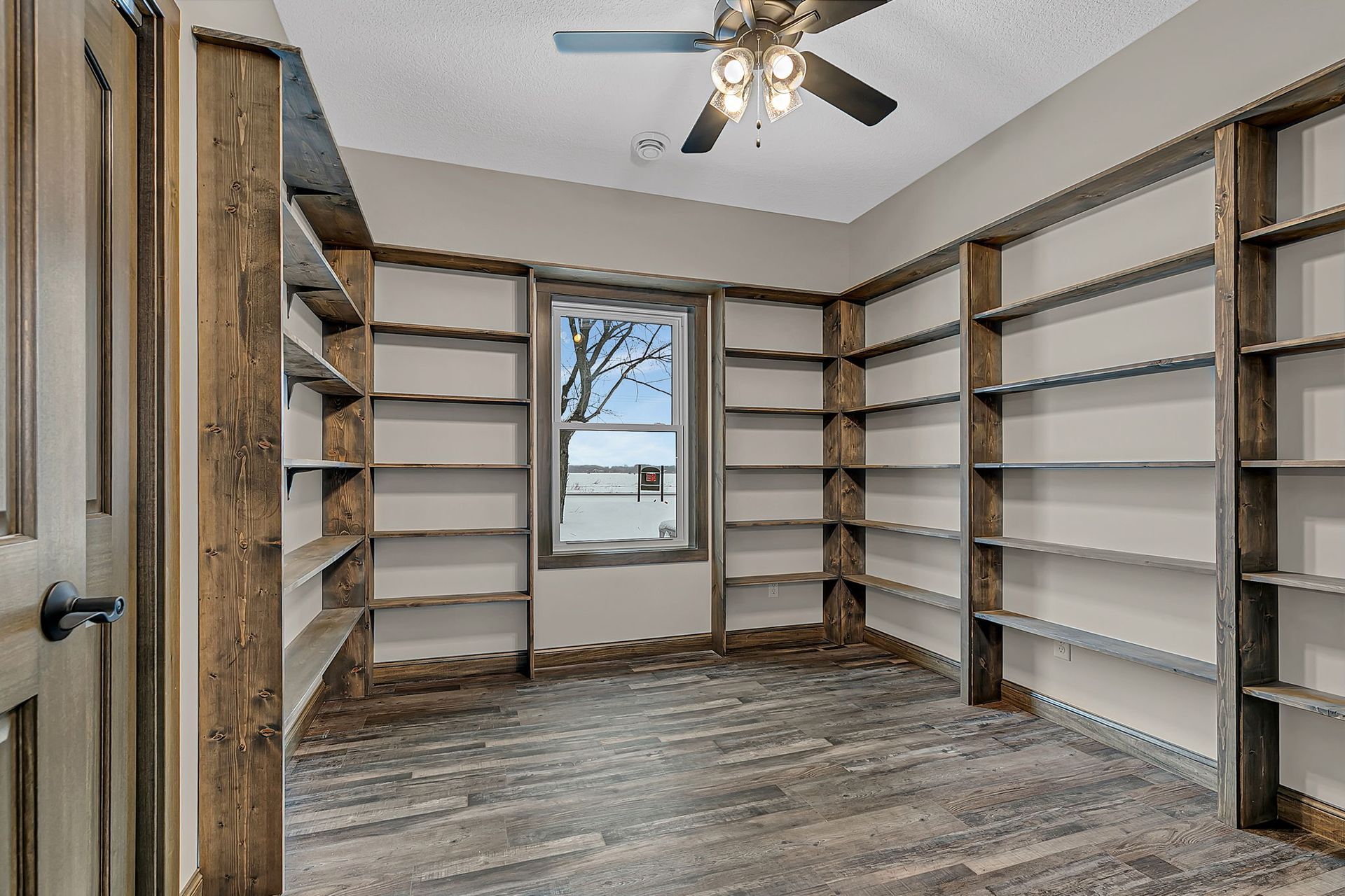 Empty room with built-in bookshelves on three walls, window, wooden floor, ceiling fan.