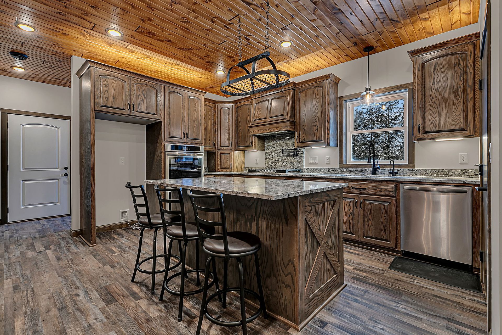 Rustic kitchen with wood cabinets, island with stools, stainless steel appliances, and wood ceiling.