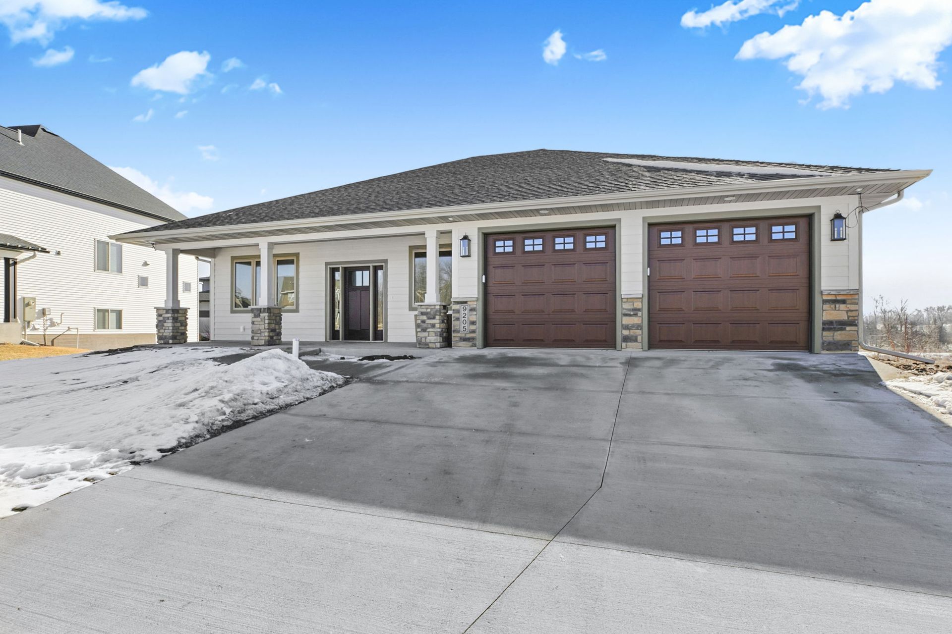 Modern beige house with a two-car garage, brown doors, and concrete driveway. Snow on the side.