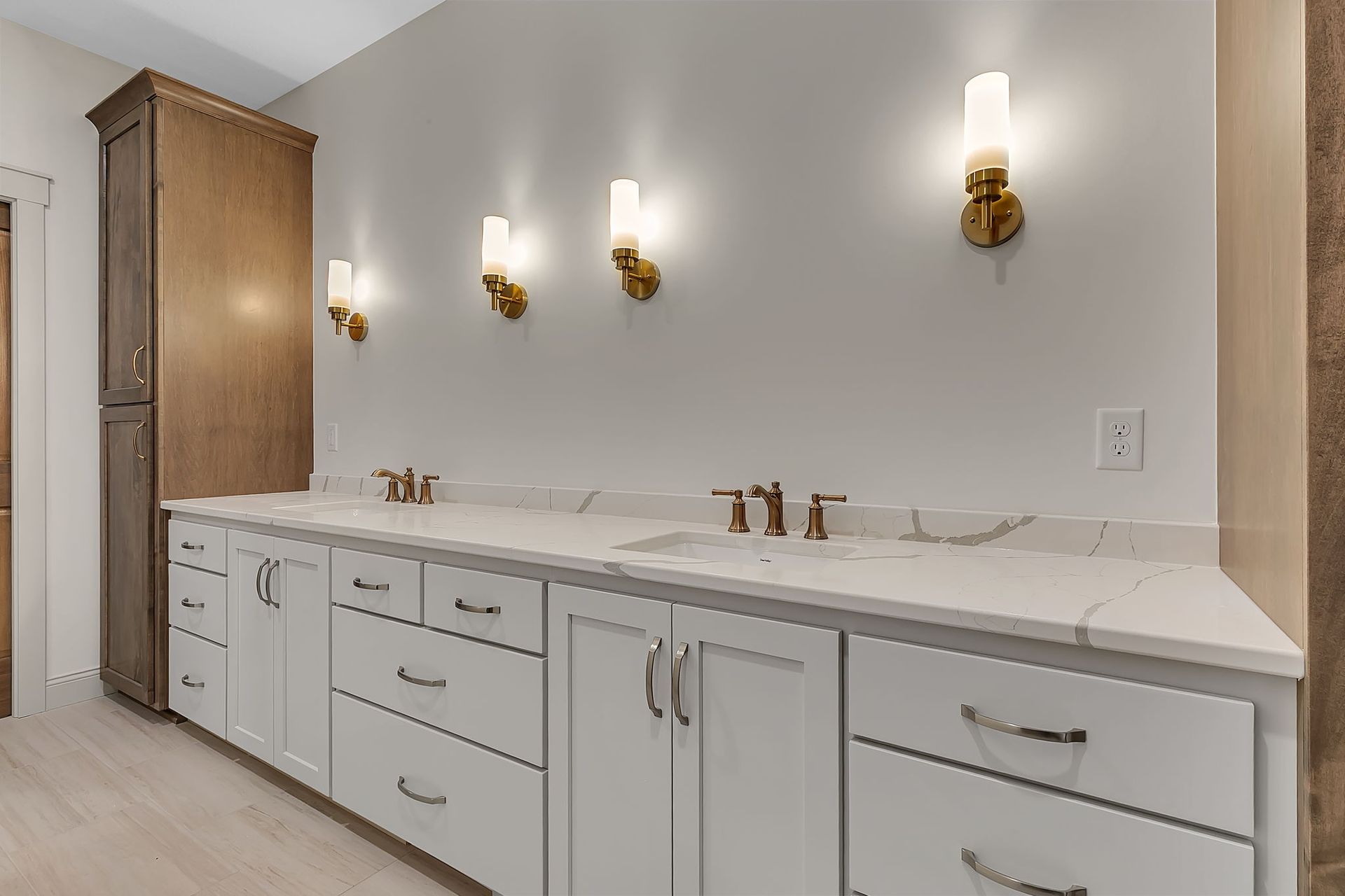 Bathroom with white vanity, quartz countertop, gold sconces, and tall wooden cabinet.