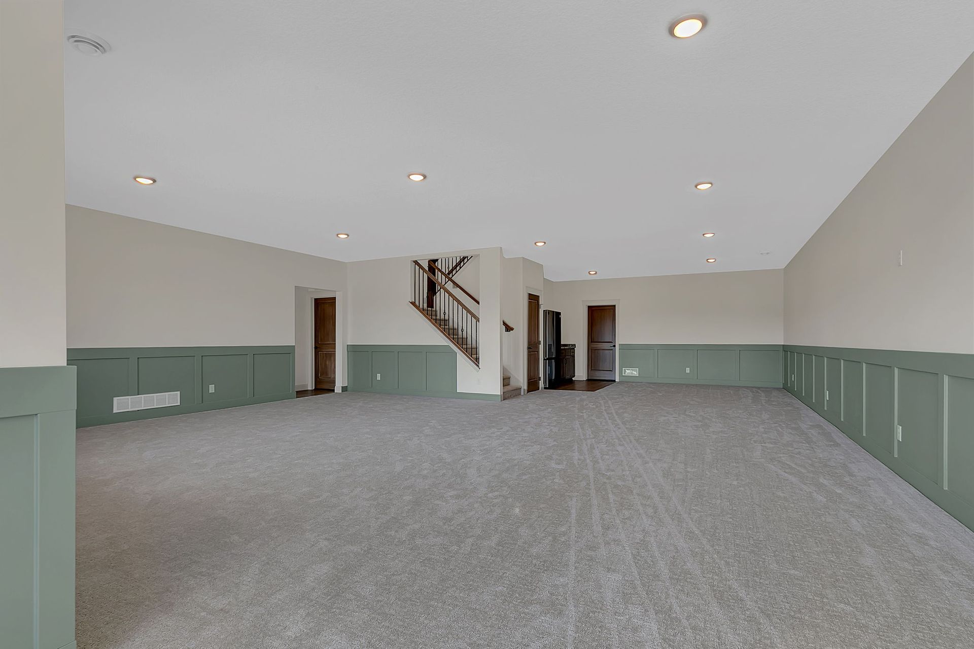Empty, carpeted room with green wainscoting and white walls, recessed lighting, and a staircase.