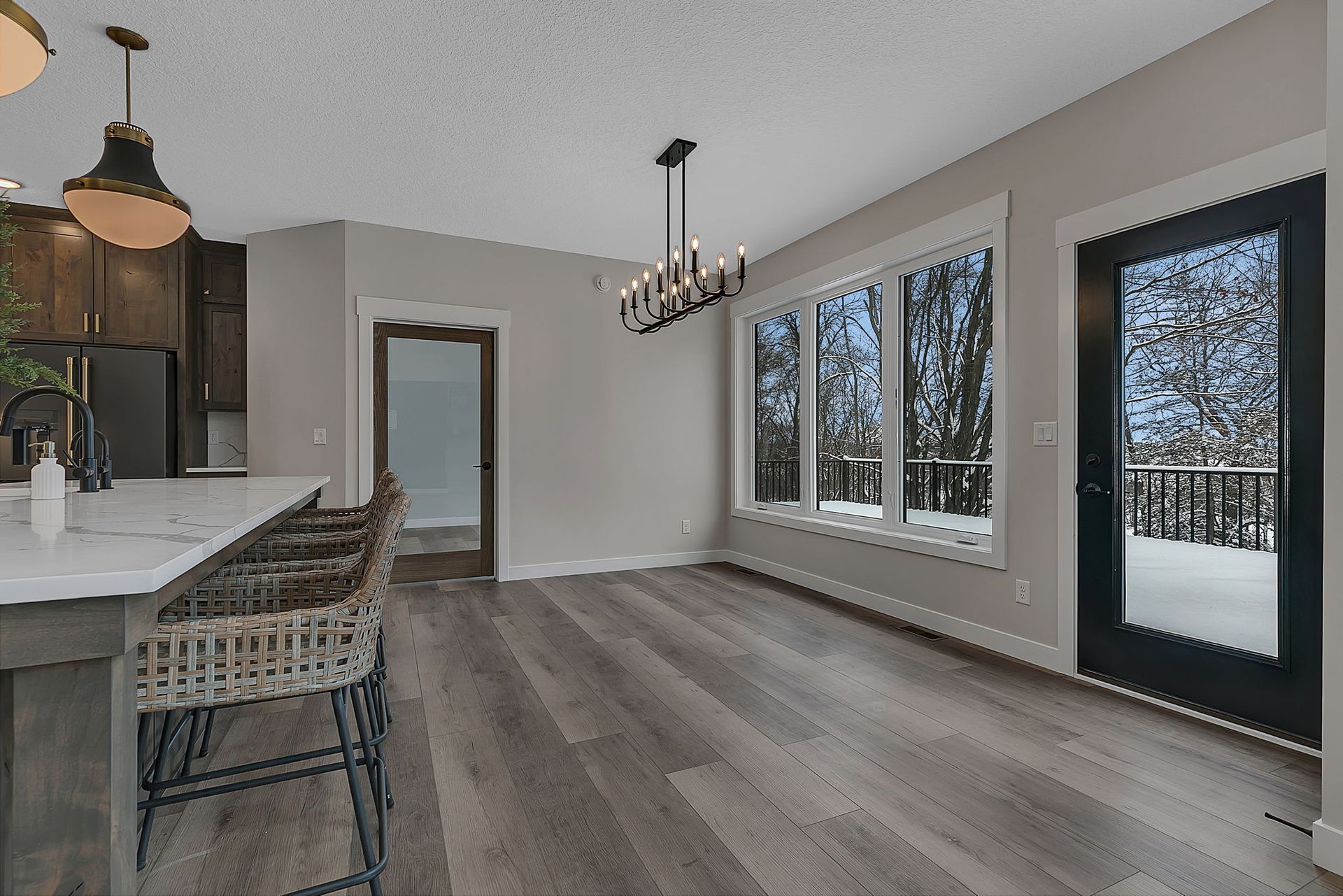 Modern dining room with large windows overlooking a snowy deck and trees. Light wood floors, neutral walls, and black trim.