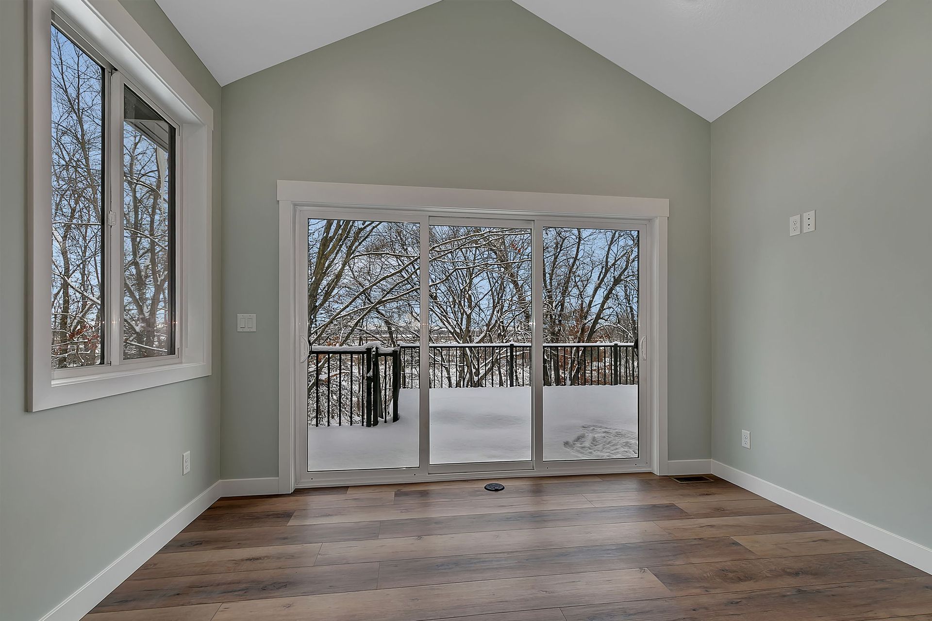 Room with large windows and sliding glass door, looking out at a snowy deck and trees.