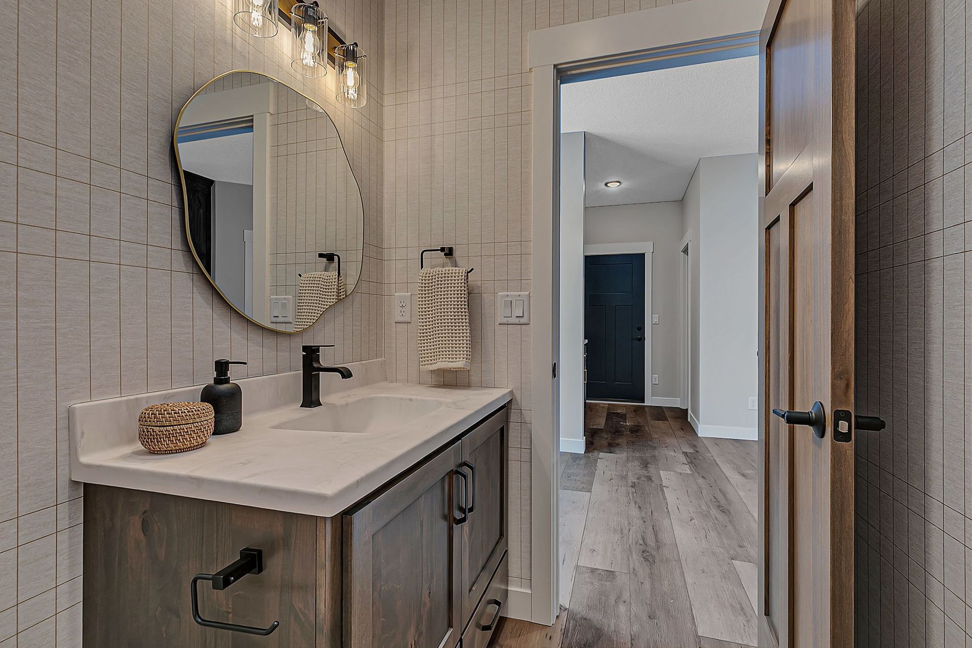 Bathroom with a wooden vanity, a unique mirror, and wood-look flooring. Doorway leads to a hallway.