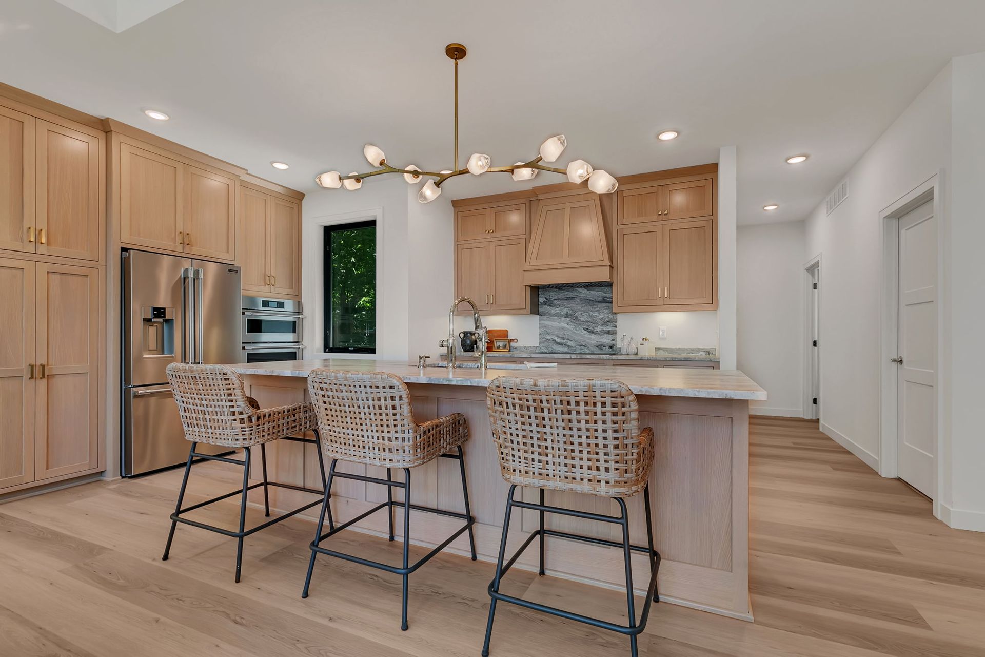 Modern kitchen with light wood cabinets, marble island, woven counter stools, and a contemporary chandelier