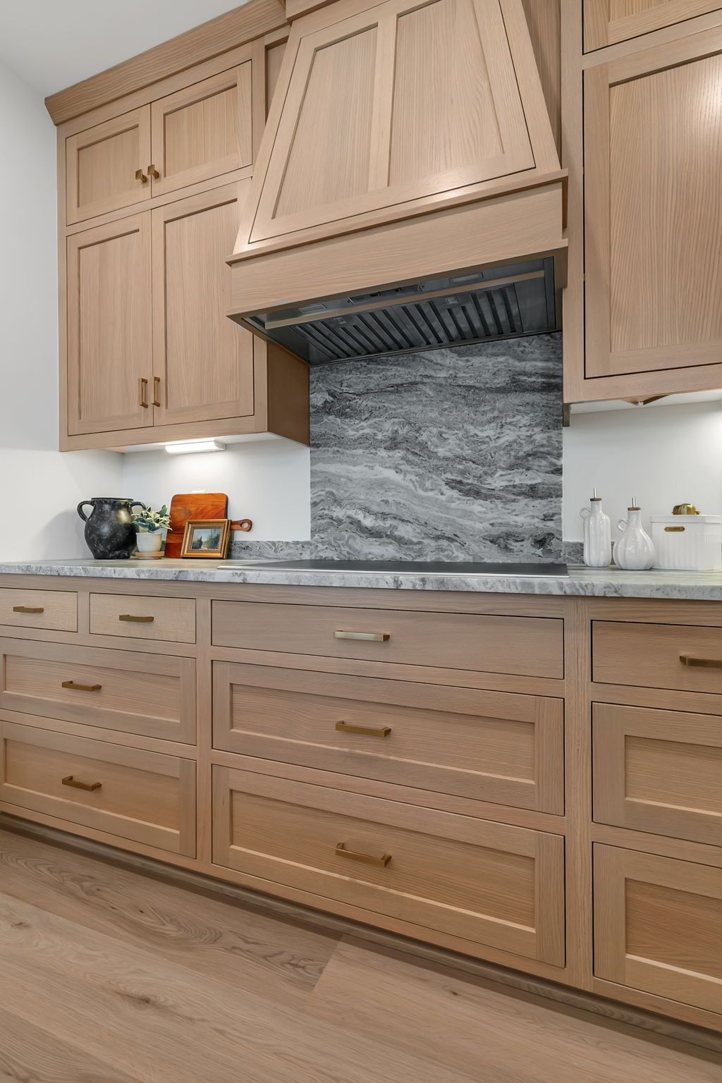 Light wood kitchen cabinets with a gray stone backsplash and matching range hood, above wood flooring