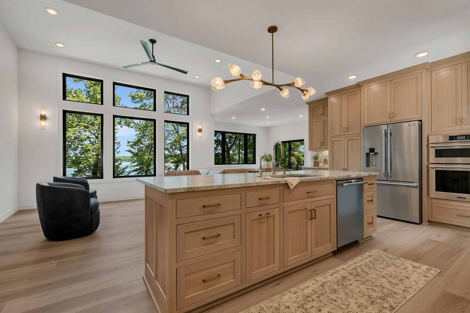 Modern open kitchen with light wood cabinets, large island, stainless steel appliances, and tall windows overlooking trees