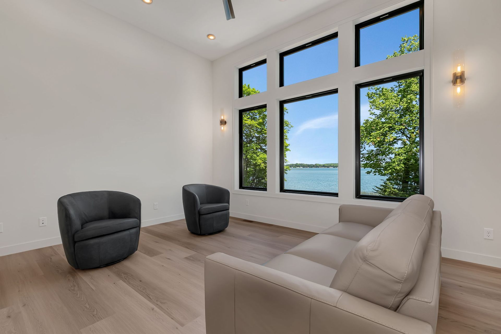 Bright living room with large windows, light wood floors, and two dark chairs facing a beige sofa.