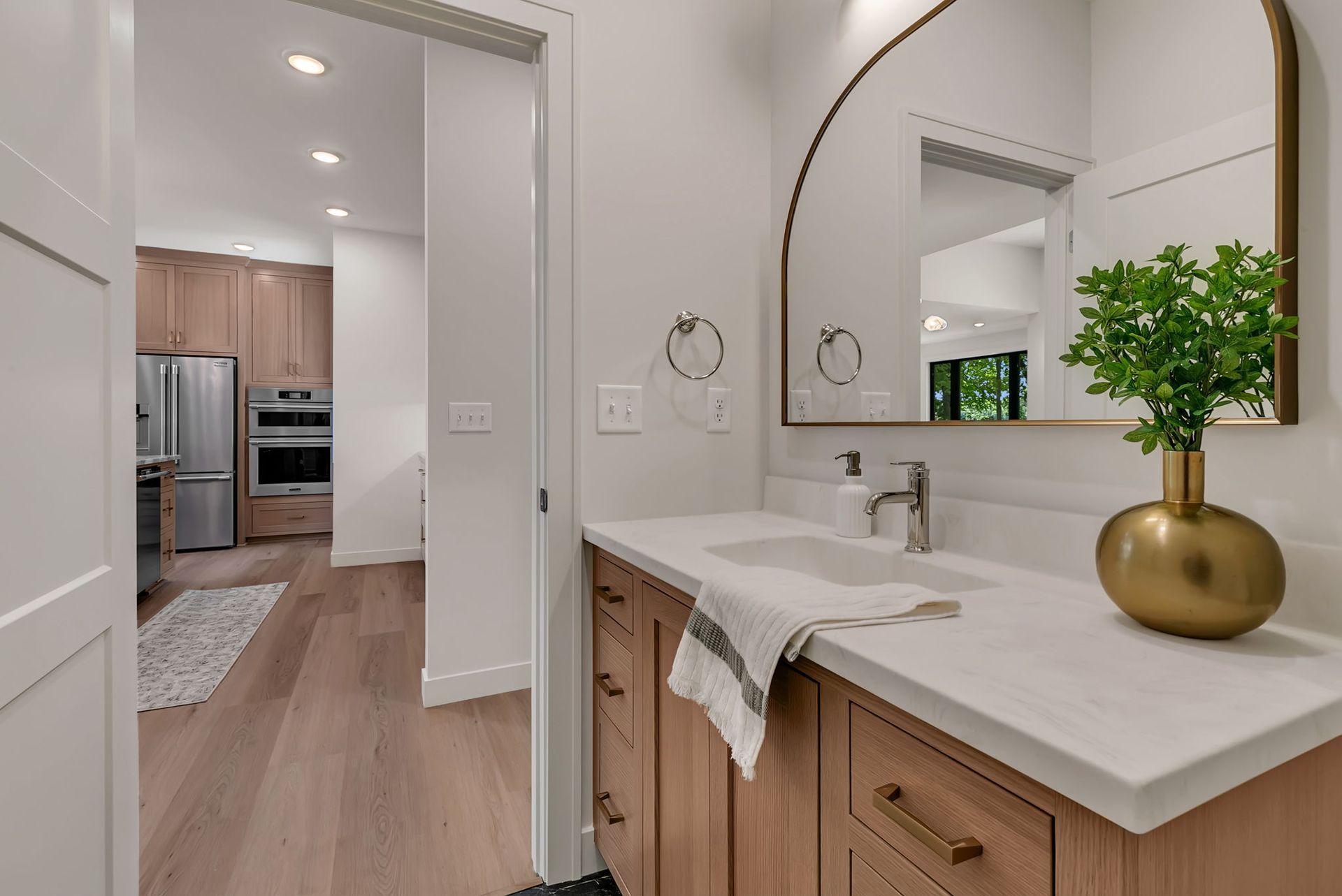 Modern bathroom vanity with arched mirror and marble countertop, opening to a bright kitchen beyond.
