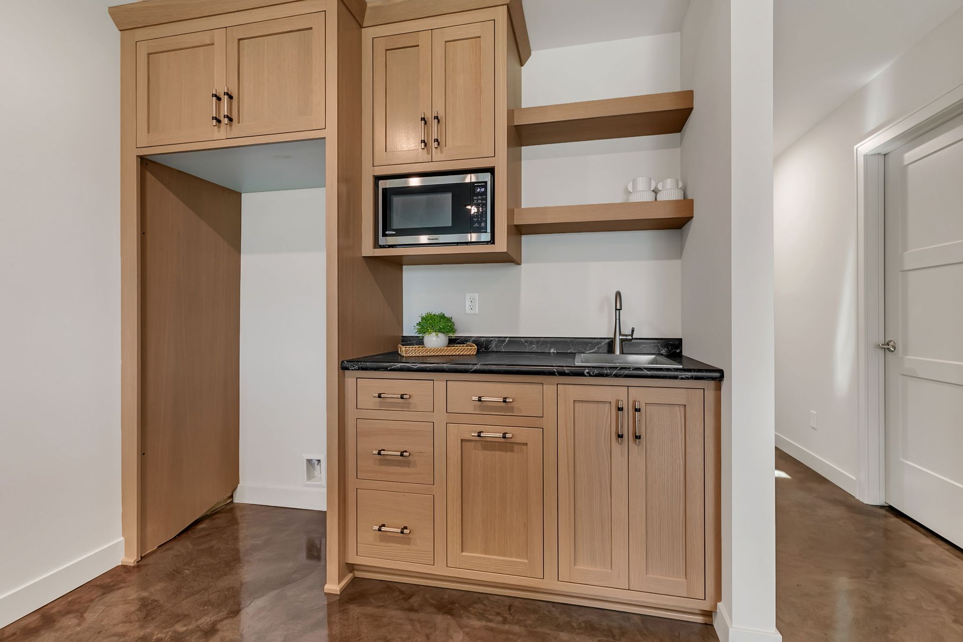 Small kitchenette with wood cabinets, microwave, sink, and open closet-like nook in a hallway.