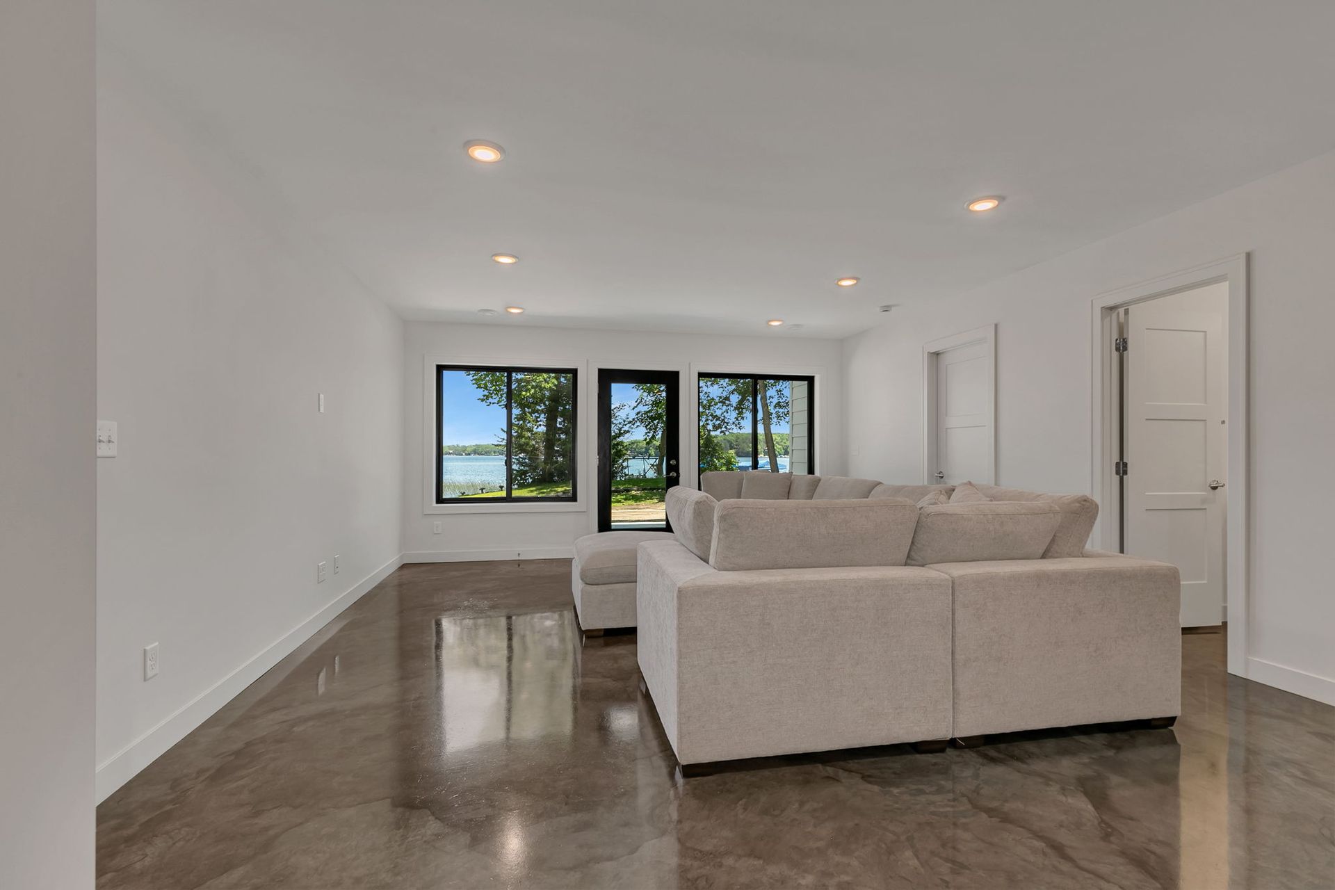 Bright empty living room with beige sectional, dark floor, and large windows at the back