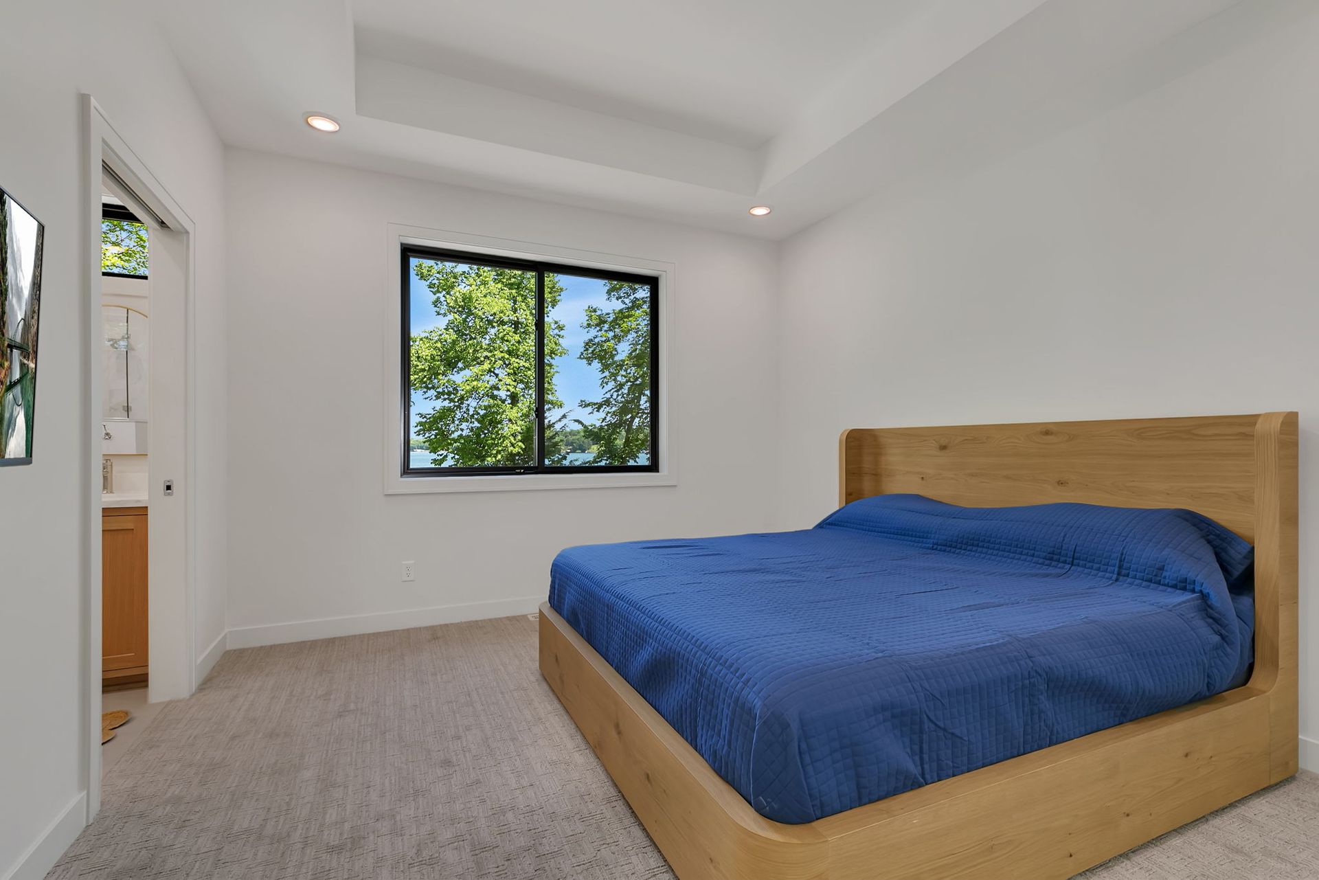 Bright bedroom with a blue bedspread, wood bed frame, and a window showing green trees.