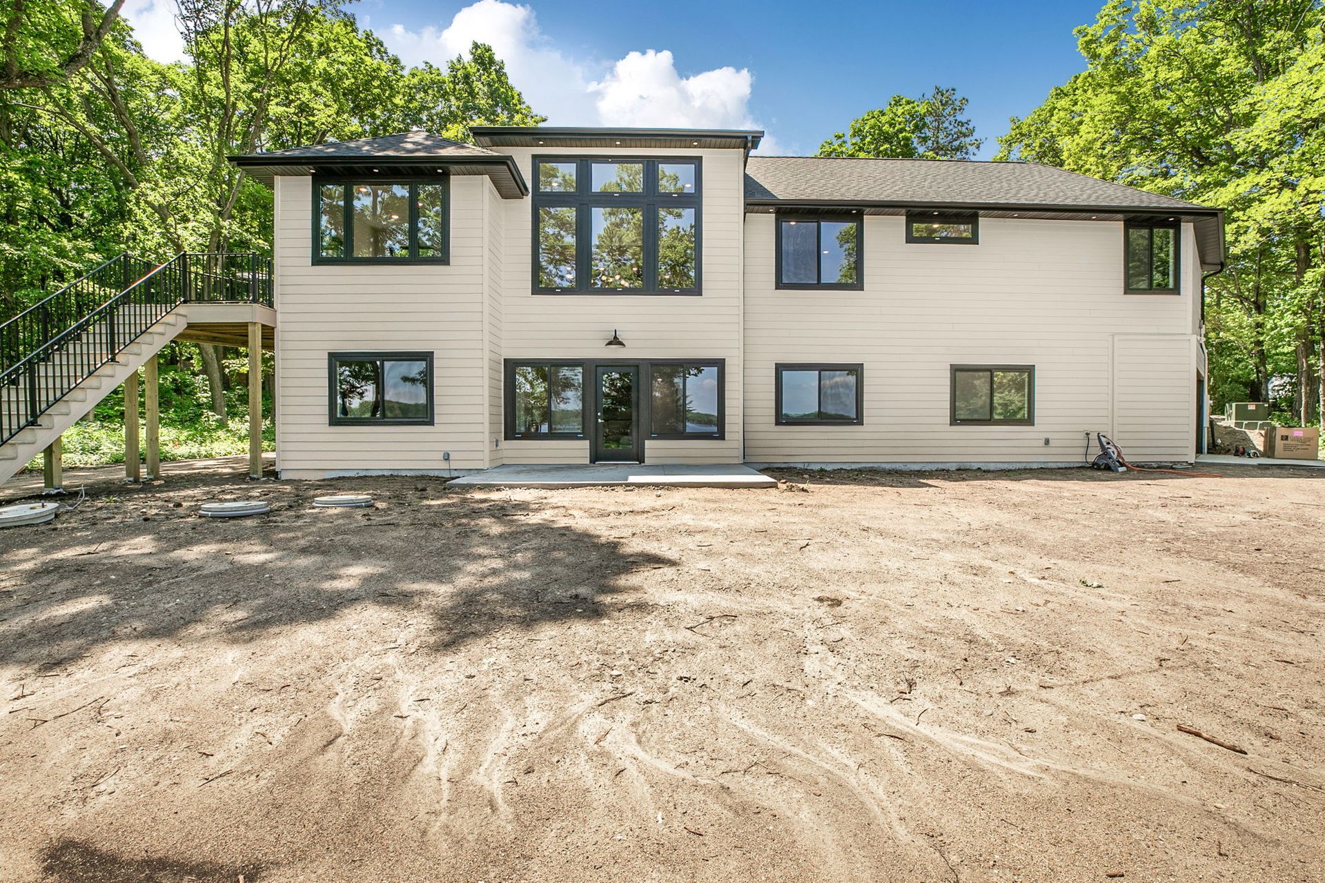 Modern two-story house with beige siding, dark trim, and a dirt front yard surrounded by trees