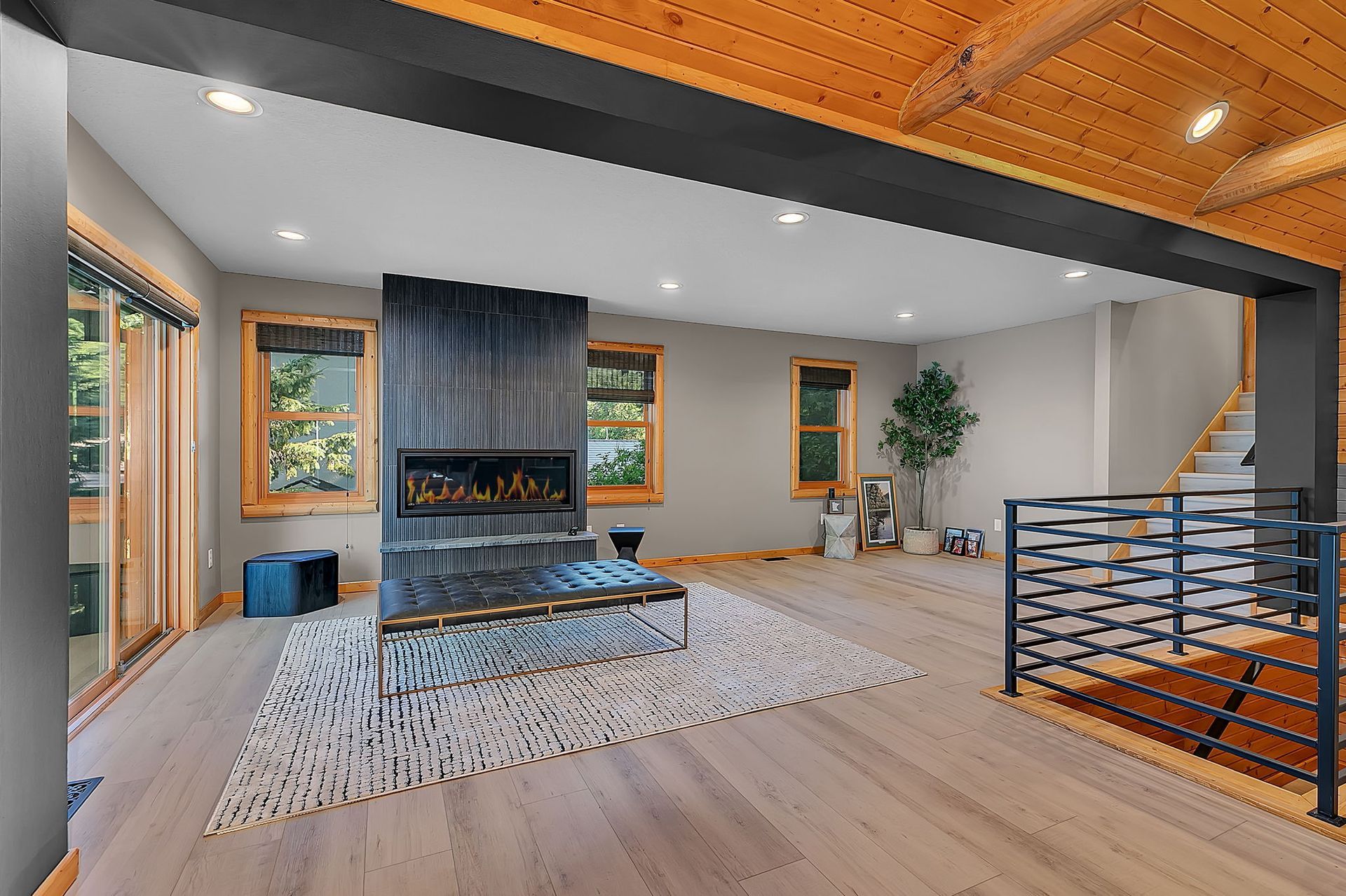 Modern living room with fireplace, gray rug, wood ceiling, stairs, and large windows