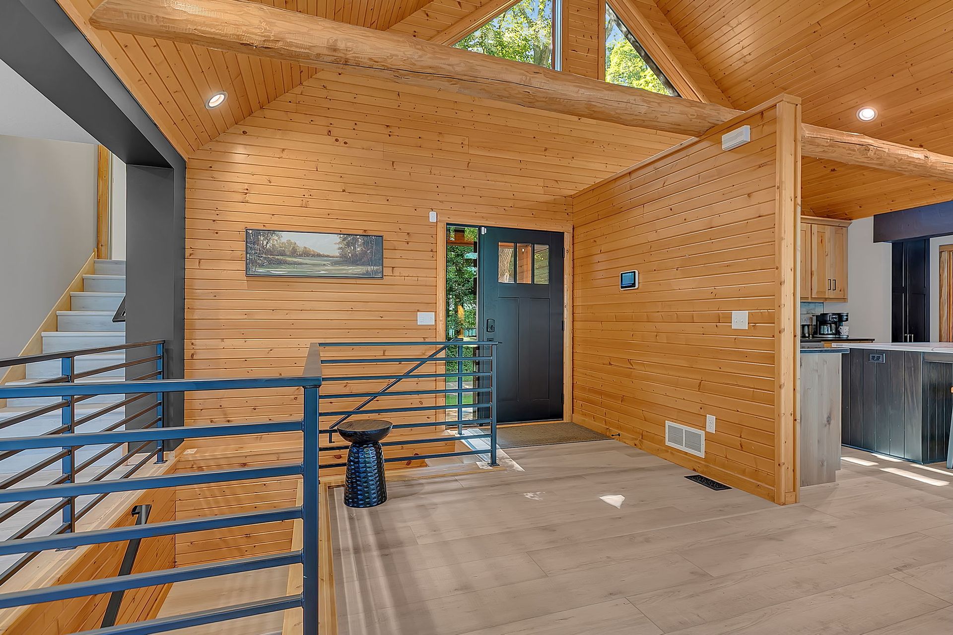 Modern wood-paneled interior with exposed beams, stairs, railing, and a blue door.