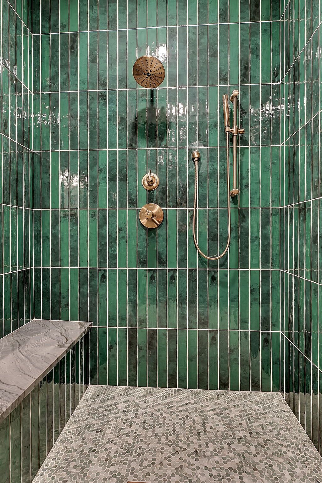 Green tiled shower with brass fixtures, handheld showerhead, and a stone bench and floor