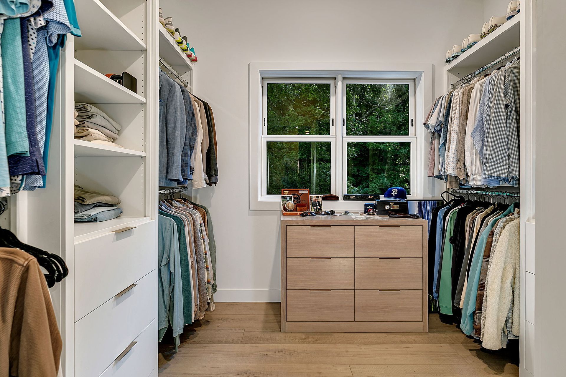 Bright walk-in closet with built-in shelves, hanging clothes, and a dresser under a window.