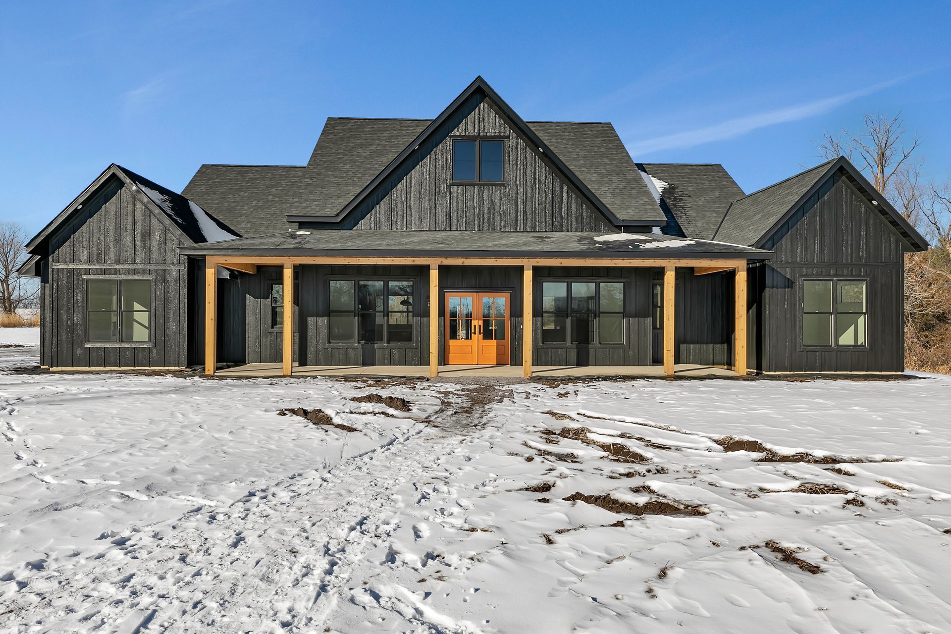 A large black house with a porch is sitting in the middle of a snowy field.