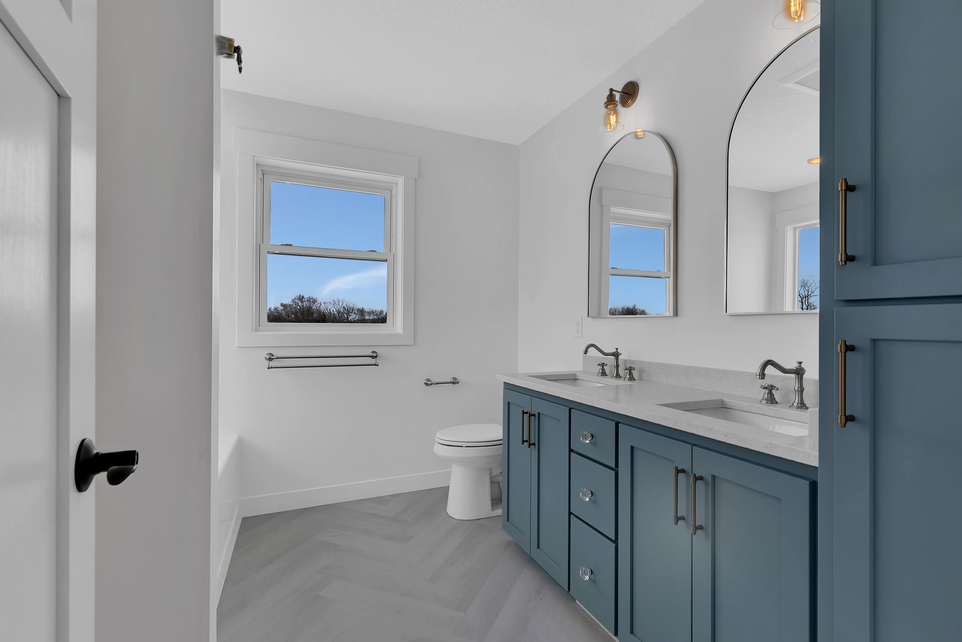 A bathroom with blue cabinets , two sinks , a toilet and two mirrors.