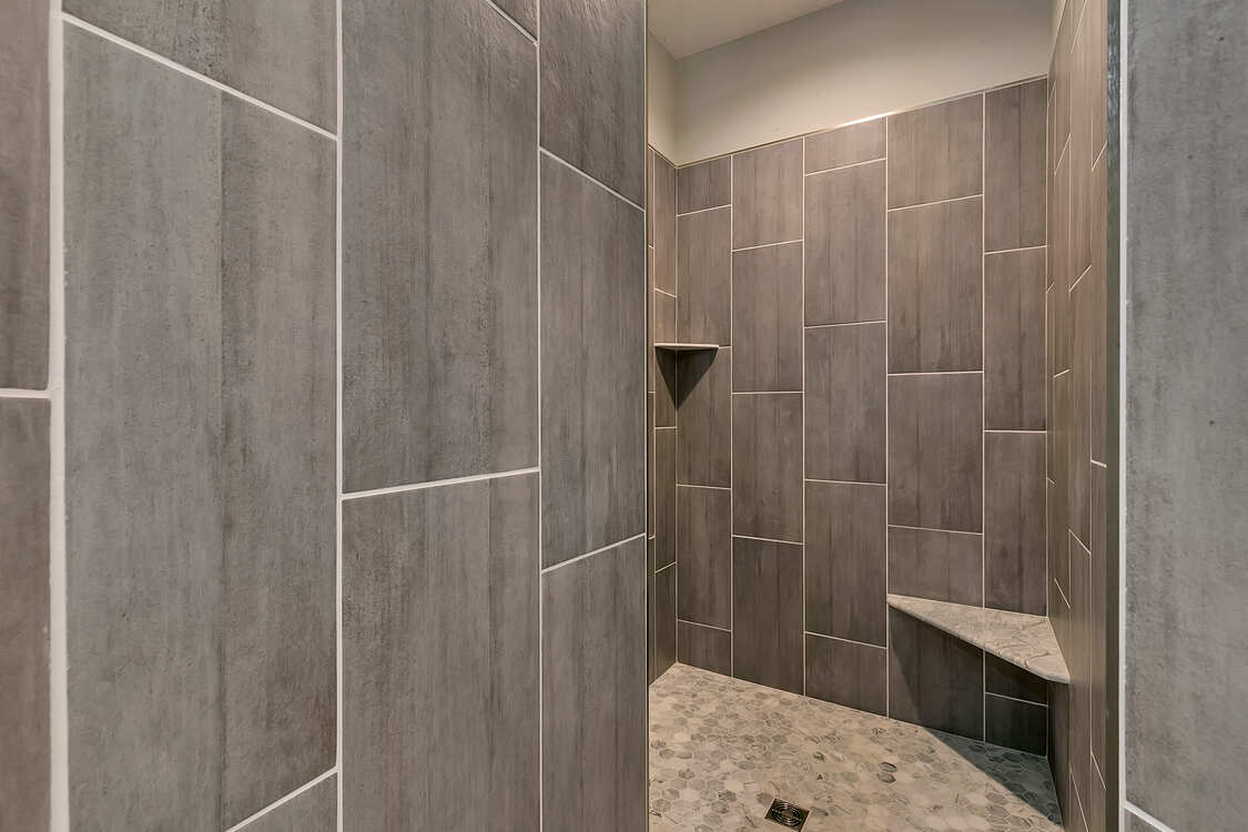 A walk in shower with gray tiles and a bench.